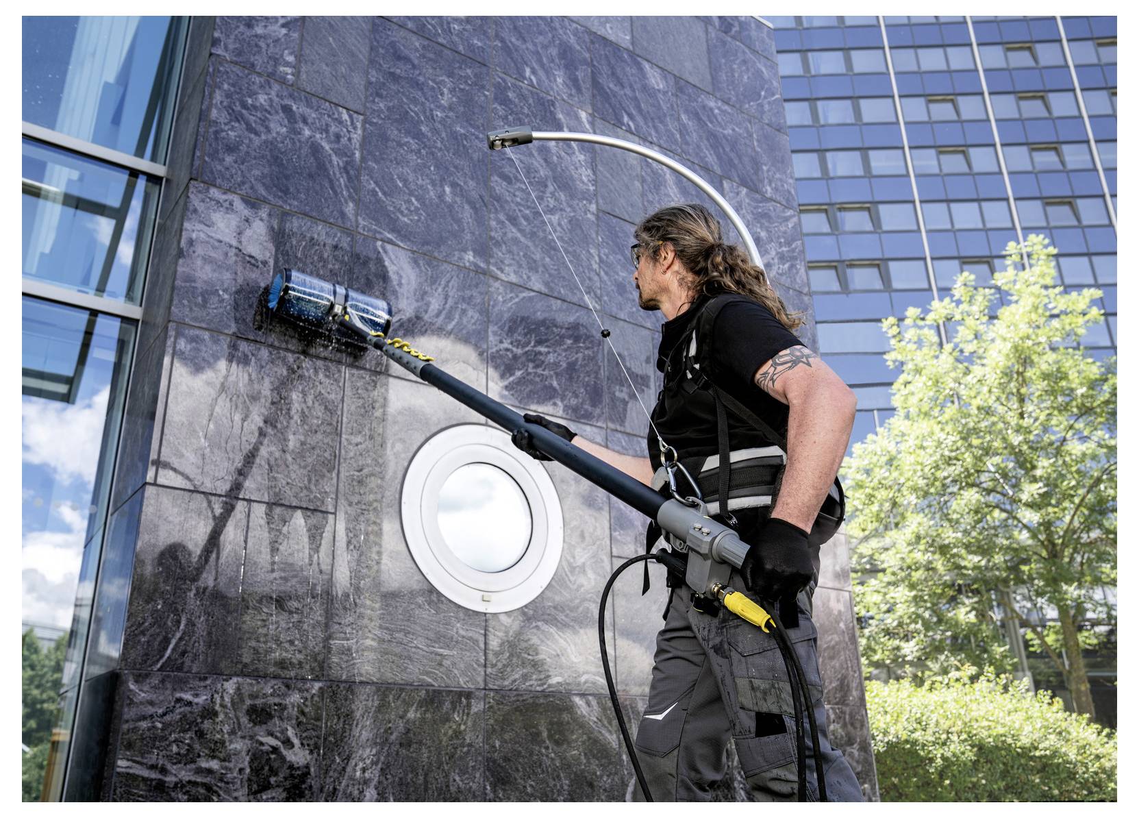 Eine Person benutzt ein Elektrowerkzeug, um die Außenseite eines Gebäudes zu reinigen, wobei sie sich auf ein rundes Fenster konzentriert, mit Bäumen und blauem Himmel im Hintergrund.