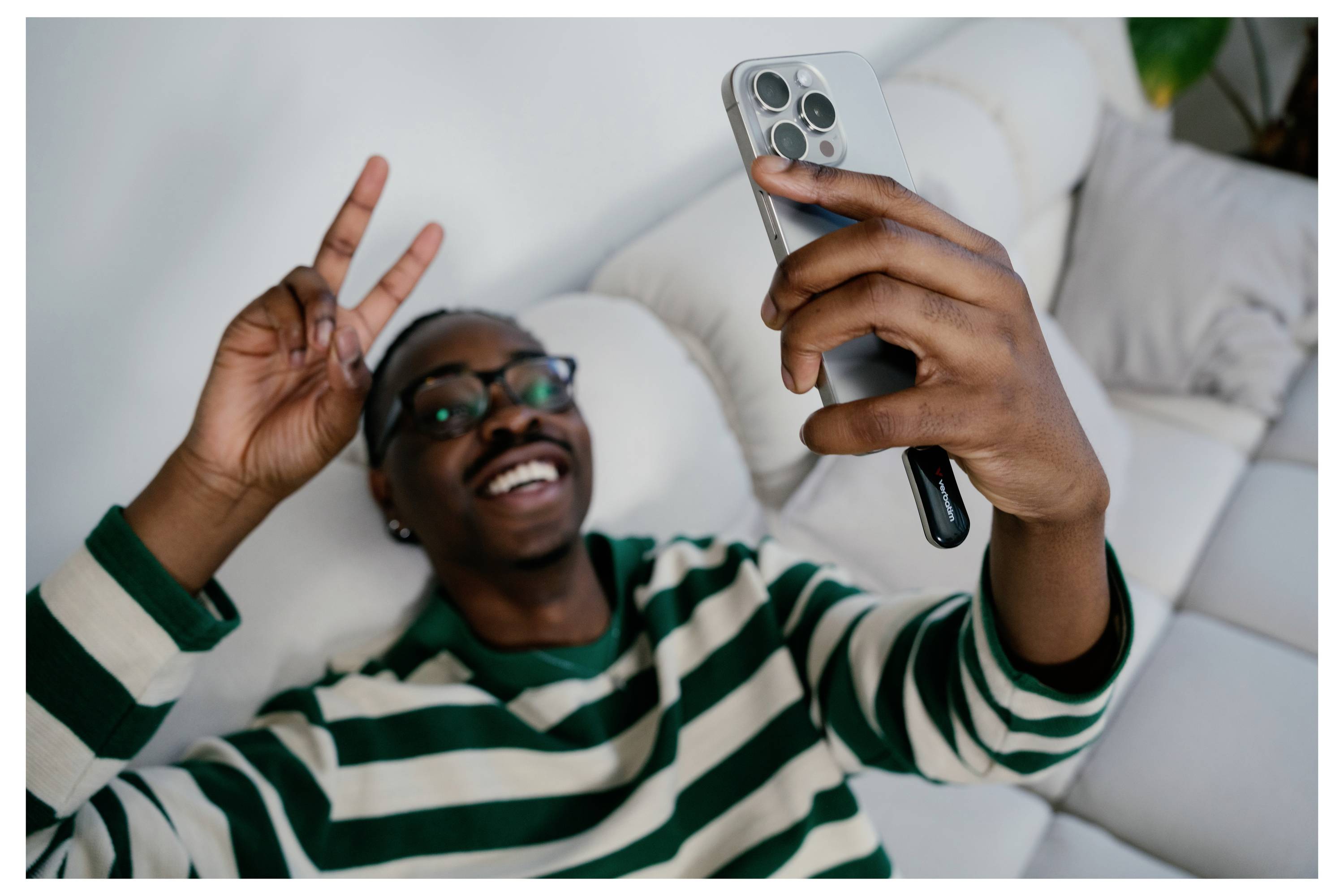 Lächelnder Mann, der auf einem Sofa liegt und ein Selfie mit einem Smartphone macht und dabei ein Friedenszeichen zeigt.