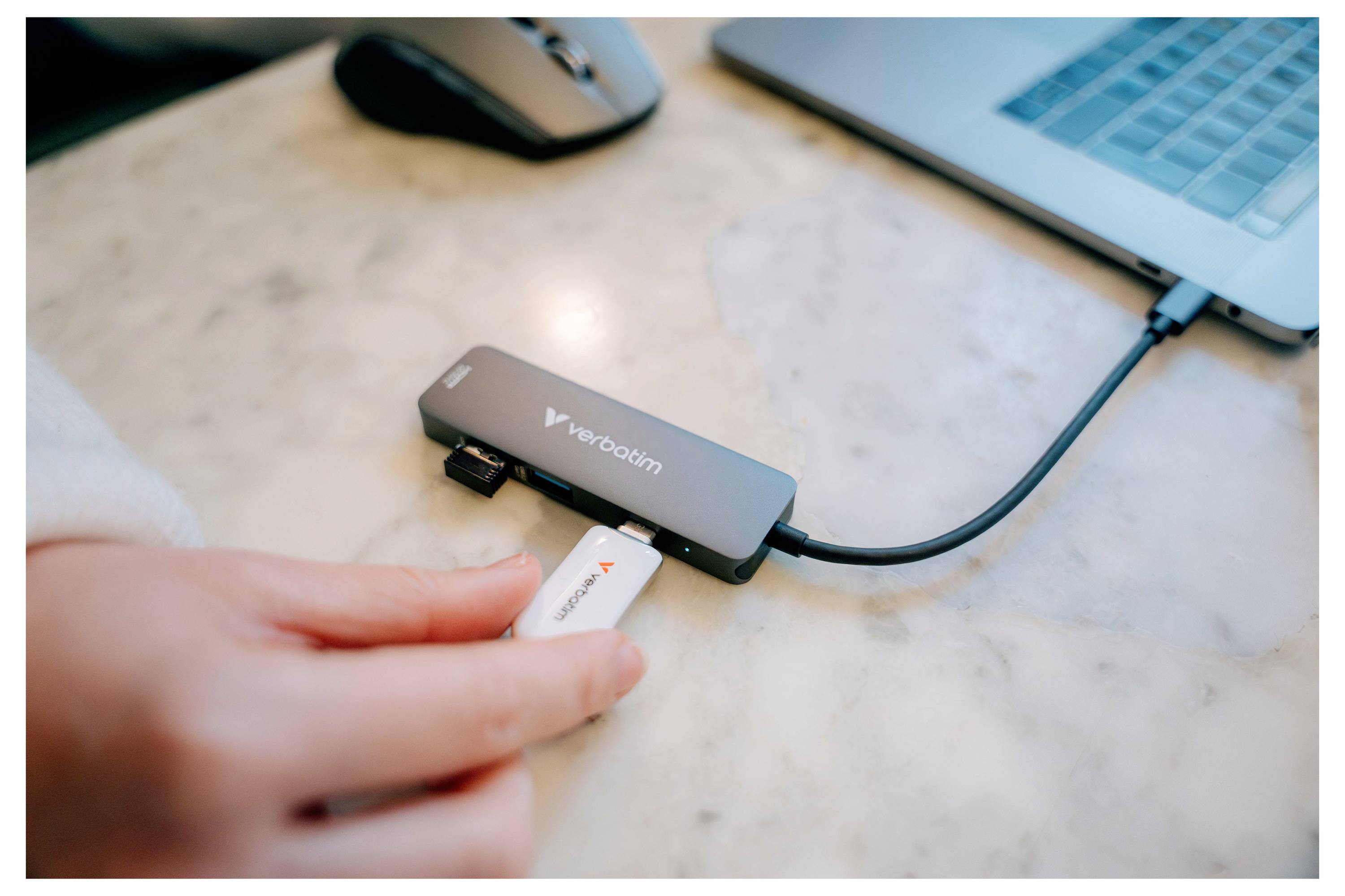 Eine Hand steckt einen USB-Stick in einen Verbatim-USB-Hub neben einem Laptop auf einem Schreibtisch.