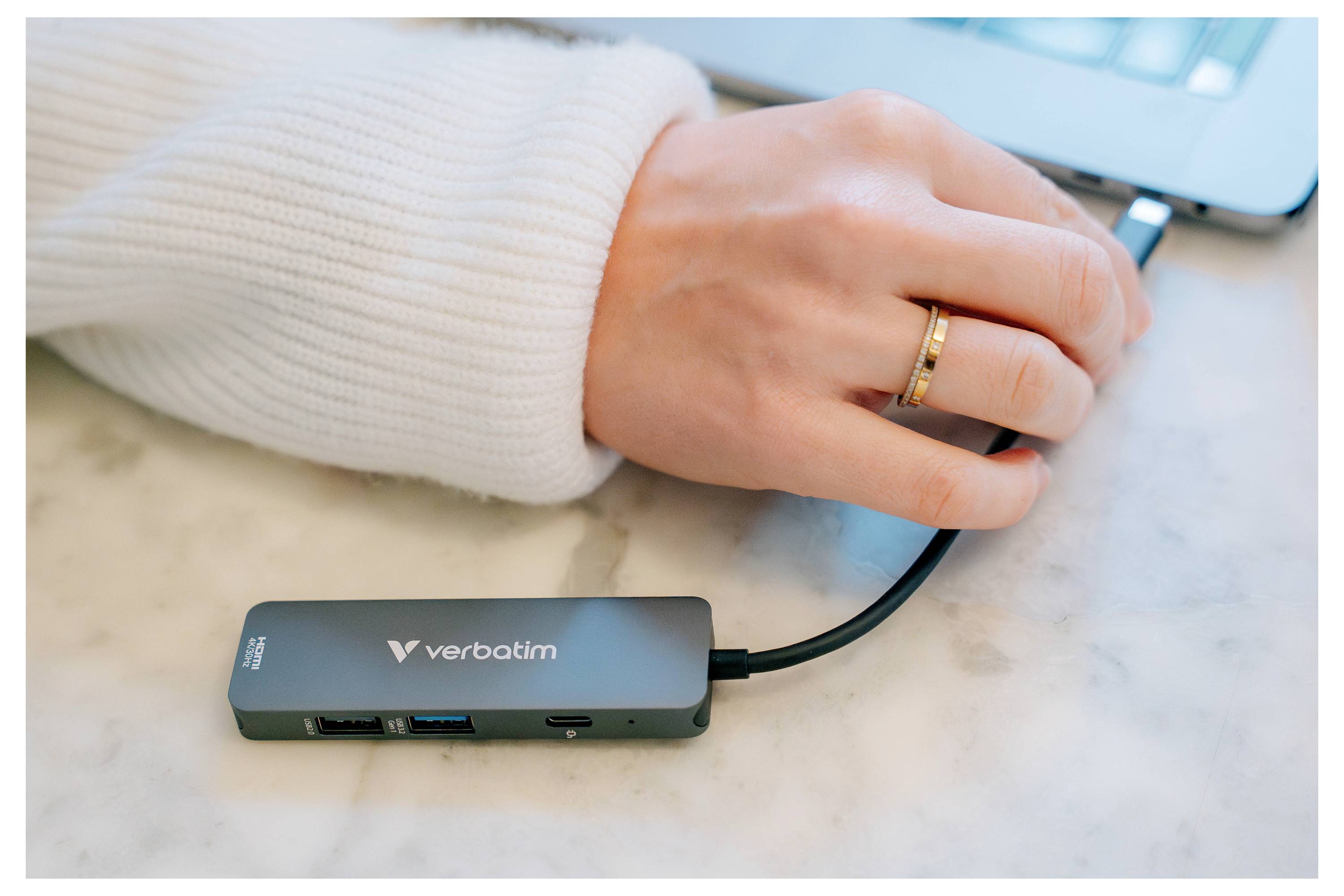 Hand, die einen Verbatim USB-C-Hub mit einem Laptop verbindet, wobei der Hub auf einer Tischplatte liegt und USB-Anschlüsse sowie das Kabel sichtbar sind.
