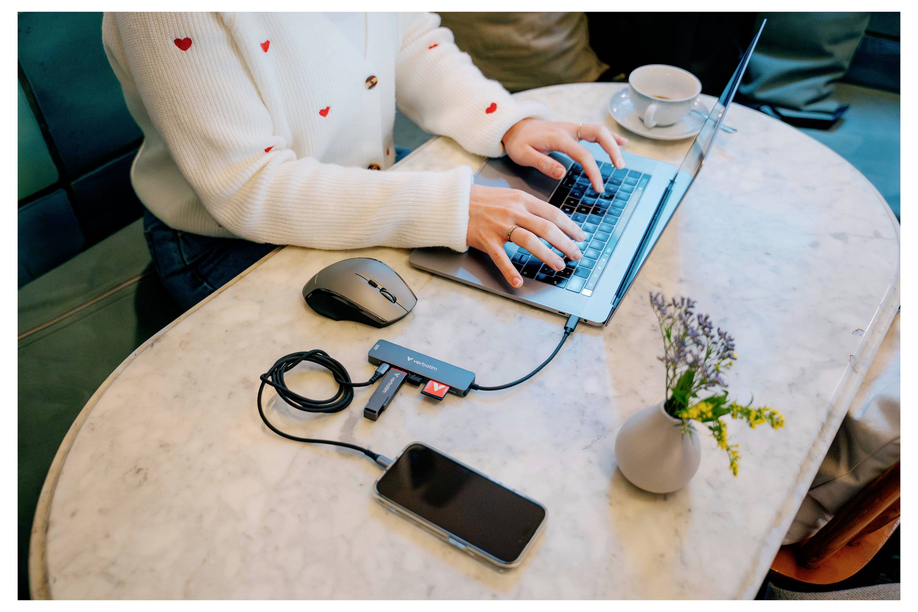 Eine Person tippt auf einem Laptop an einem Tisch mit einer Maus, einem Telefon und einem kleinen externen Laufwerk; eine Tasse und eine Vase mit Blumen stehen in der Nähe.