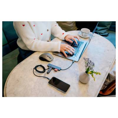 Eine Person tippt auf einem Laptop an einem Tisch mit einer Maus, einem Telefon und einem kleinen externen Laufwerk; eine Tasse und eine Vase mit Blumen stehen in der Nähe.