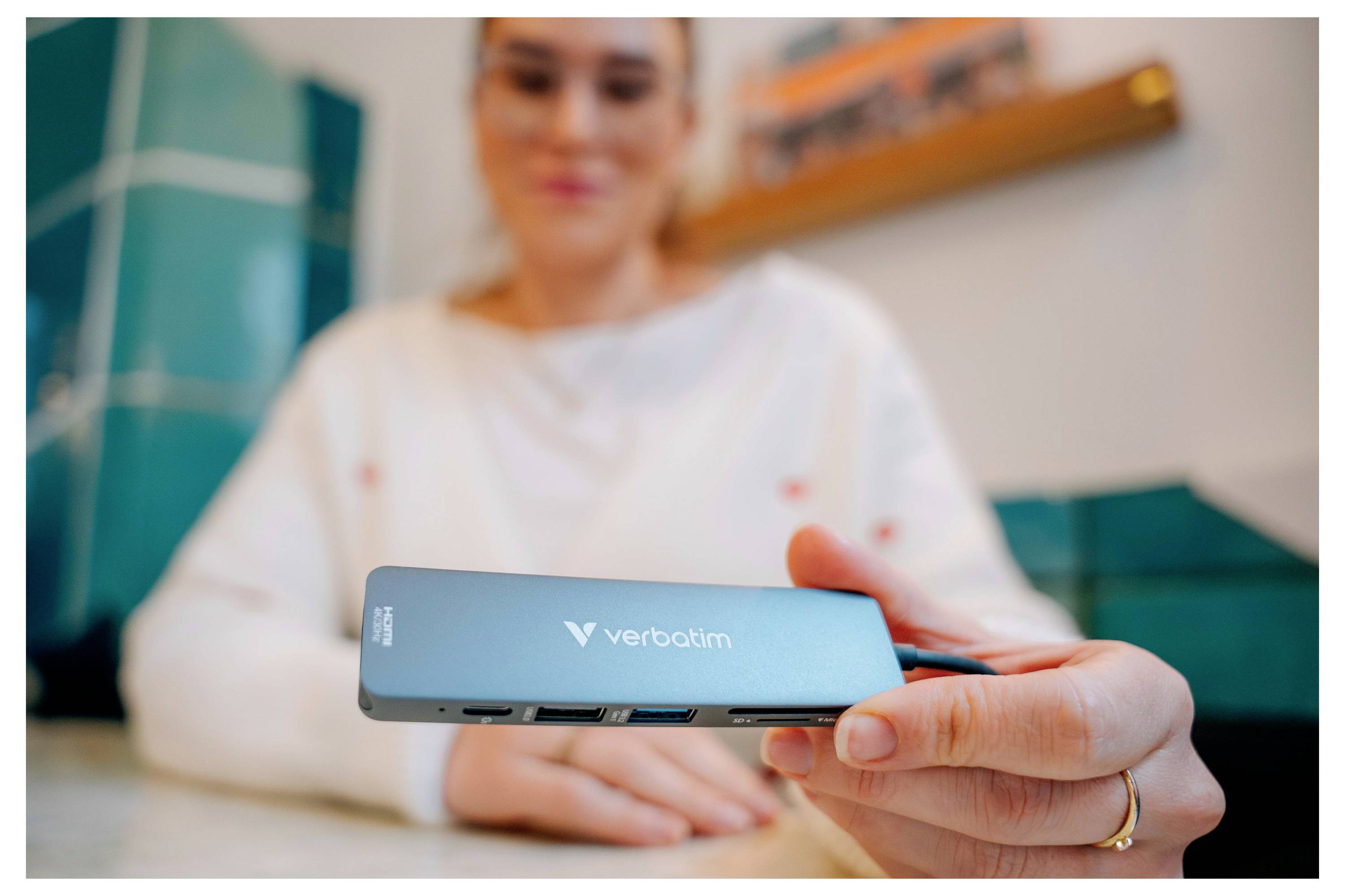 Eine Frau hält einen Verbatim USB-C-Hub in die Höhe und zeigt seine Anschlüsse im Vordergrund, während sie drinnen sitzt.