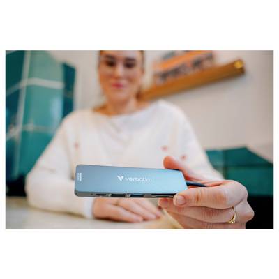Eine Frau hält einen Verbatim USB-C-Hub in die Höhe und zeigt seine Anschlüsse im Vordergrund, während sie drinnen sitzt.