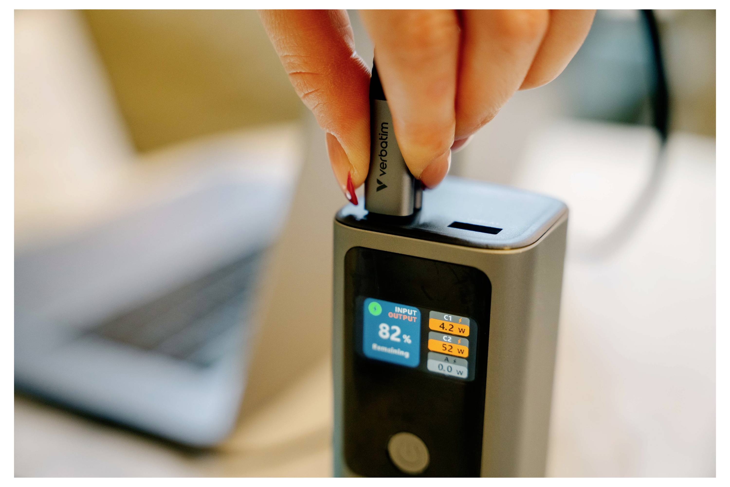 Eine Hand führt ein VapePod-ähnliches Gerät in eine schwarze Ladestation ein; ein kleiner Bildschirm zeigt 82% Batteriestand mit Ladestatistiken an.