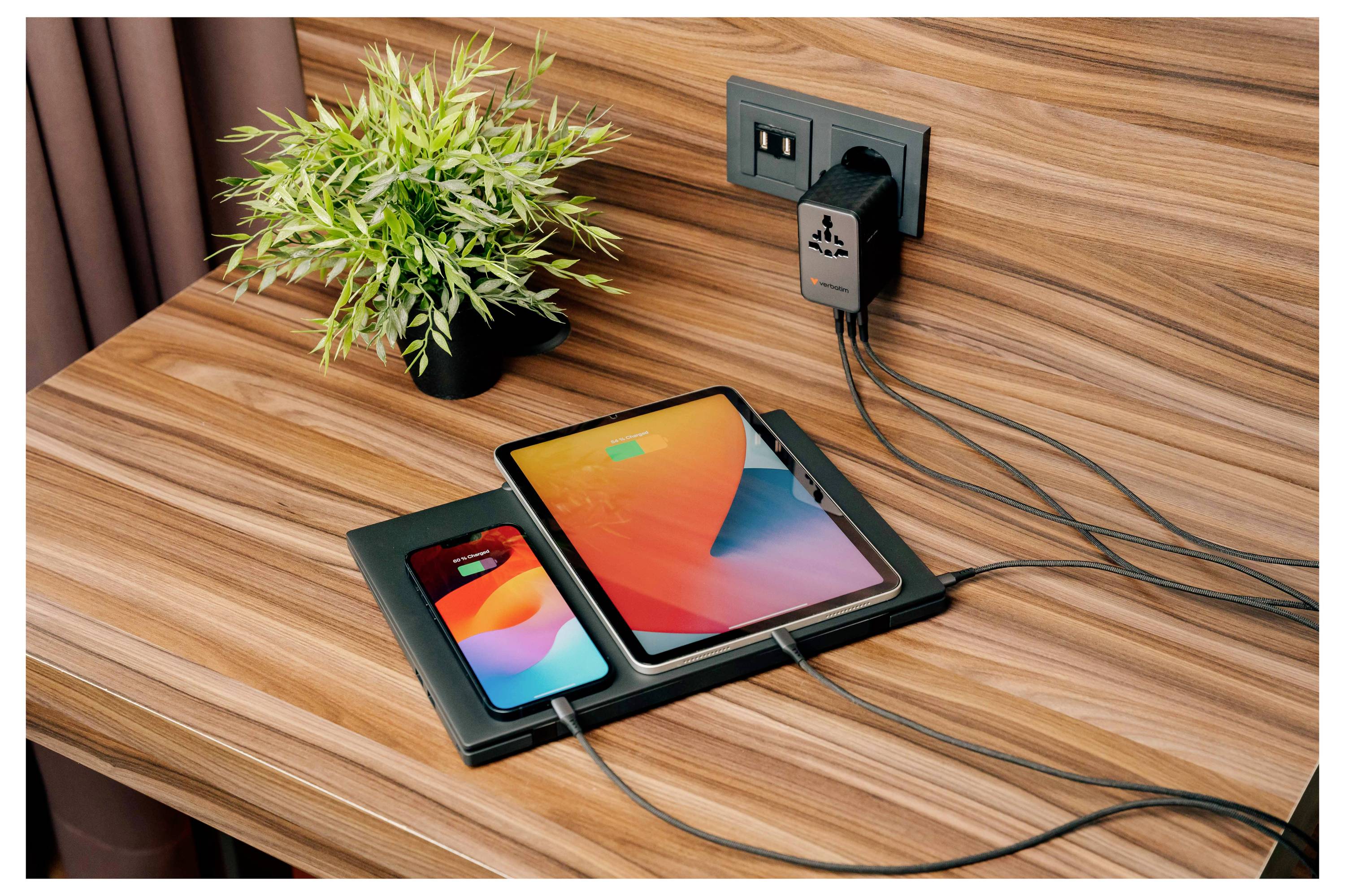 Telefon und Tablet auf einem Schreibtisch mit einem Wandadapter und Kabeln werden geladen, neben einer kleinen Topfpflanze.
