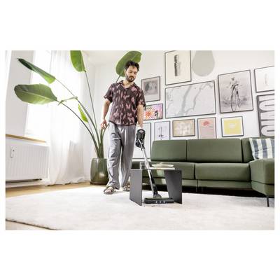 Eine Person in legerer Kleidung staubsaugt ein modernes Wohnzimmer mit einem grünen Sofa, Wandkunst und einer großen Pflanze und vermittelt damit eine entspannte Reinigungsroutine.