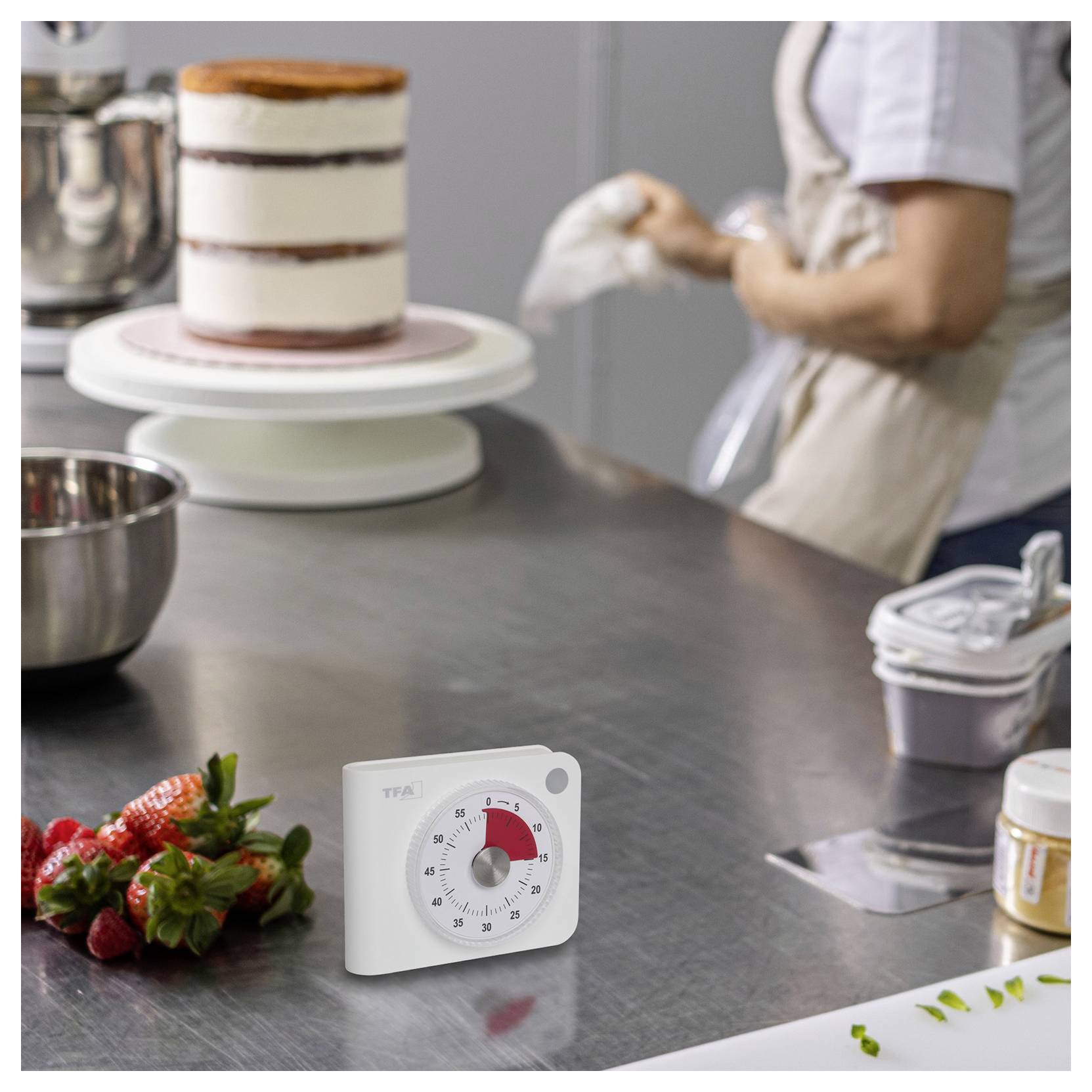 Eine Person dekoriert einen Kuchen auf einem Drehteller in einer Küche, mit Erdbeeren auf der Arbeitsplatte und einem Timer, der 10 Minuten verbleibend anzeigt.