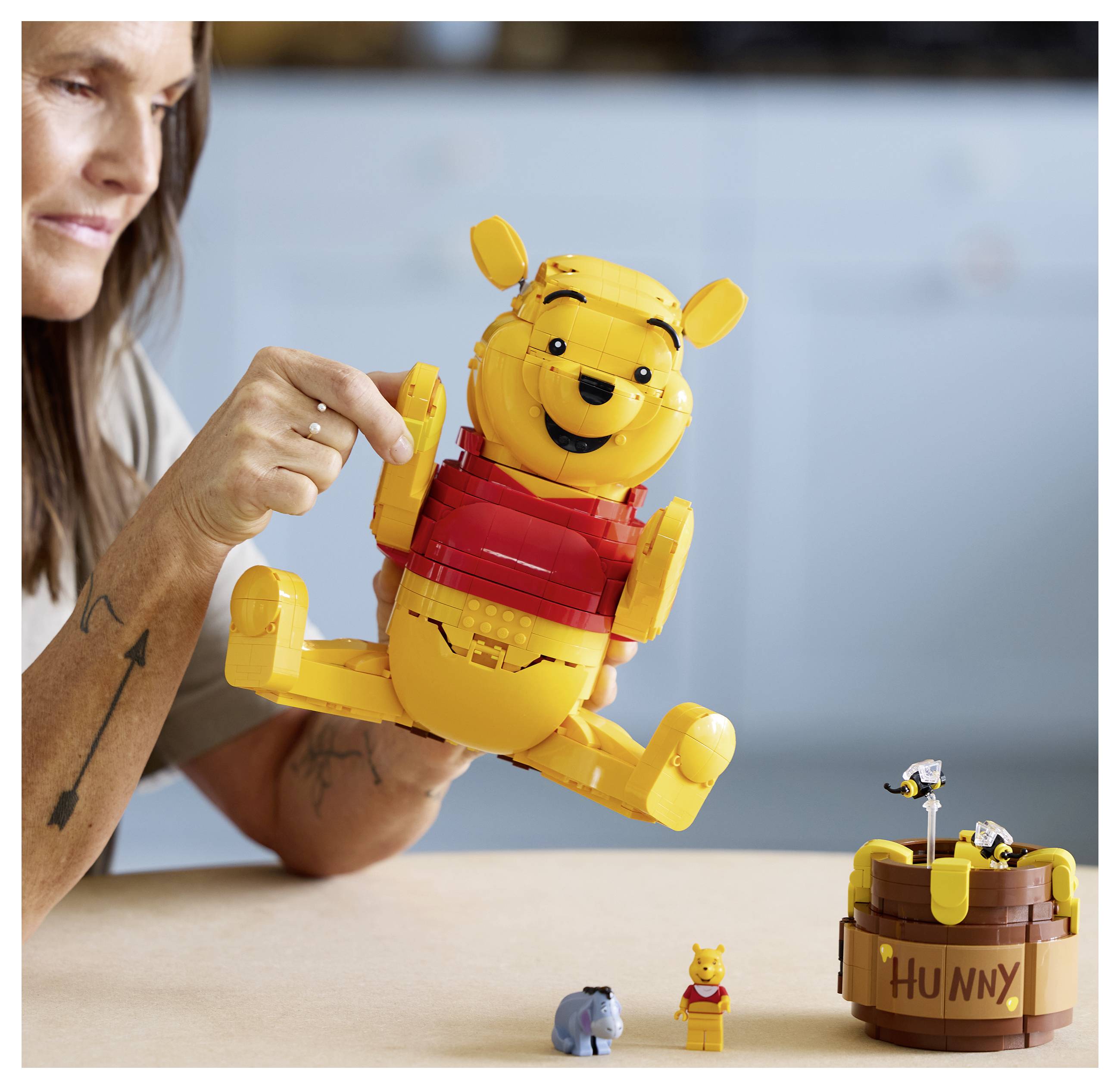 Eine Person interagiert mit einer großen, zusammengebauten Spielfigur, die einem Bären in Rot ähnelt. In der Nähe befinden sich kleinere Bären- und Eselspielzeuge sowie ein „Hunny"-Topf.