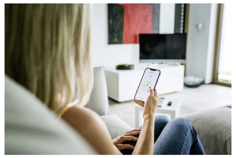 Frau sitzt auf einer Couch und benutzt ein Smartphone, mit einem Wohnzimmer und Fernseher im Hintergrund.