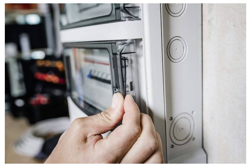Hand öffnet eine Abdeckung des Elektropanels, um auf die Schutzschalter in einem an der Wand montierten Sicherungskasten zuzugreifen.