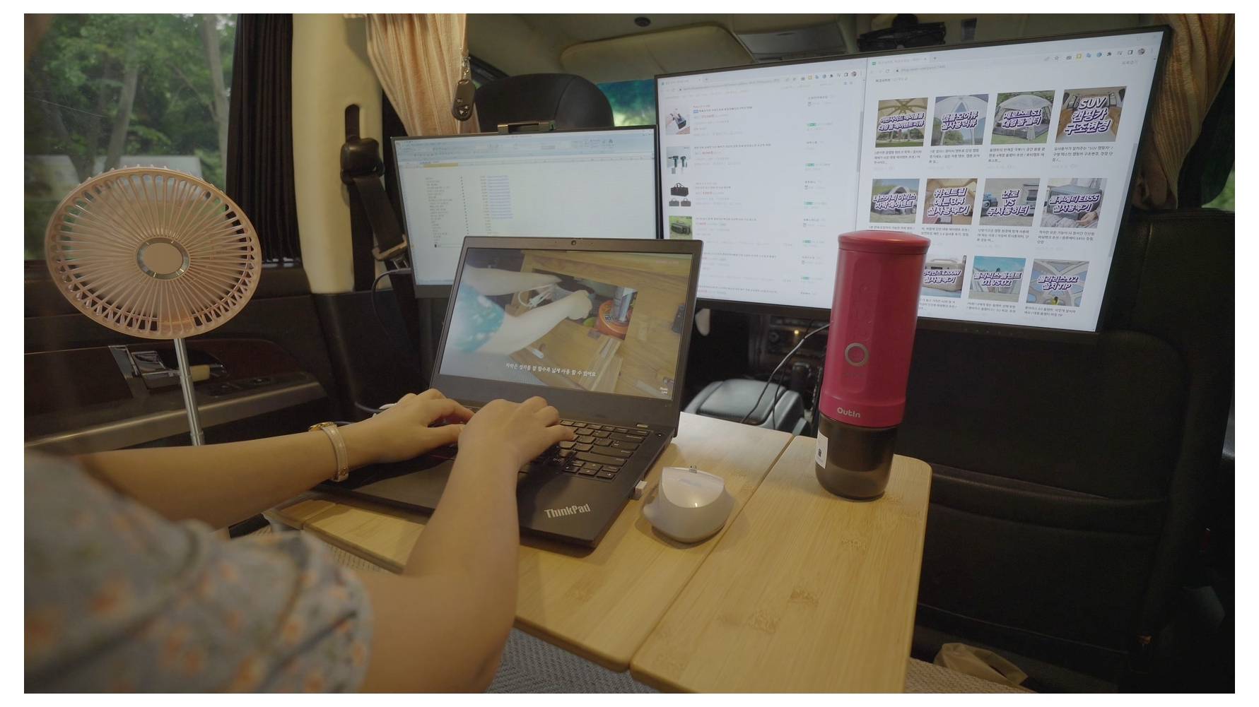 Eine Person arbeitet an einem Laptop an einem Schreibtisch in einem Fahrzeug, wobei zwei große Monitore digitale Inhalte anzeigen und ein Ventilator in der Nähe steht.
