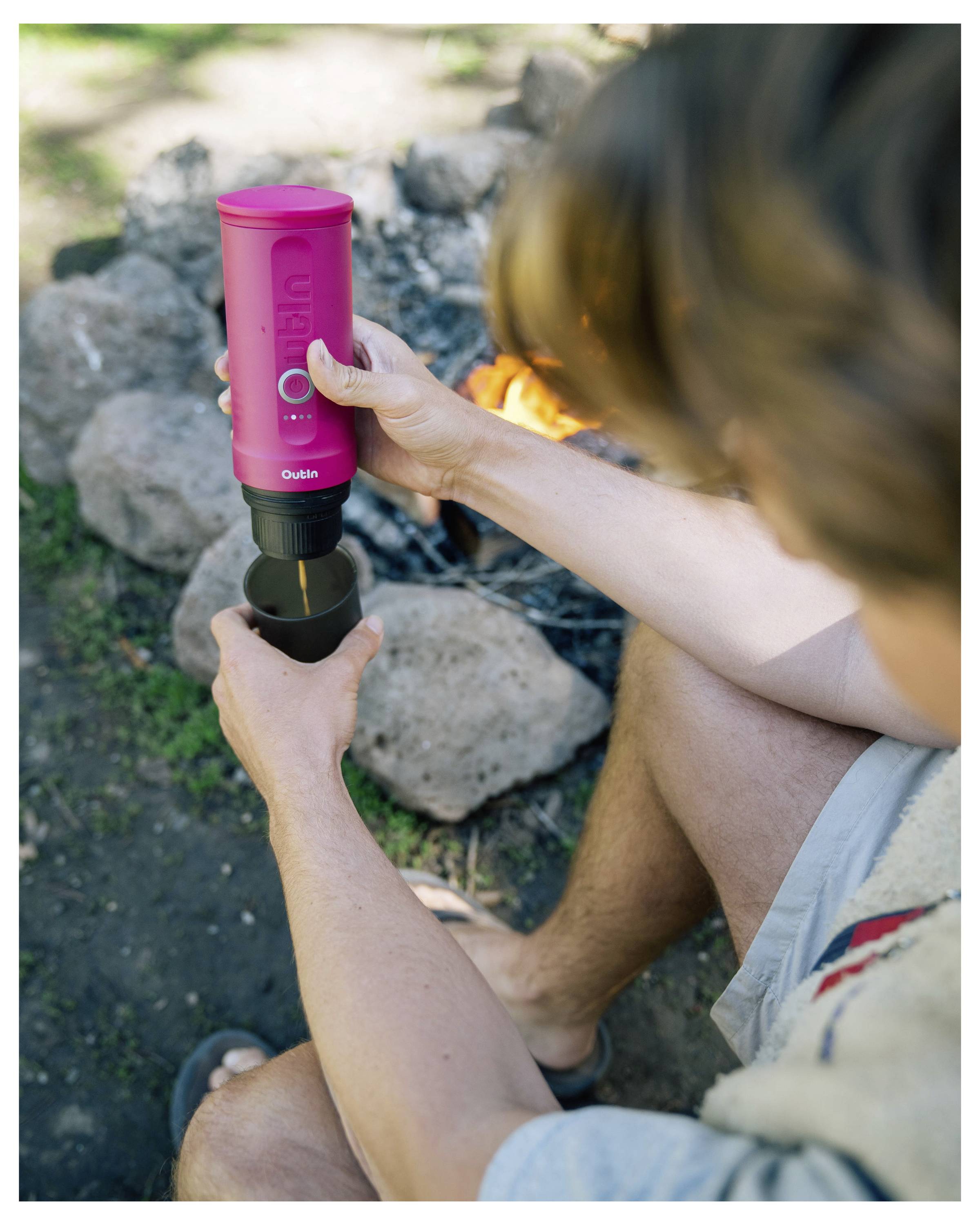 Person sitzt draußen neben einem Lagerfeuer und nutzt eine rote tragbare Kaffeemaschine, um Kaffee in eine kleine Tasse zu brühen. Geeignet für Camping-Setups.