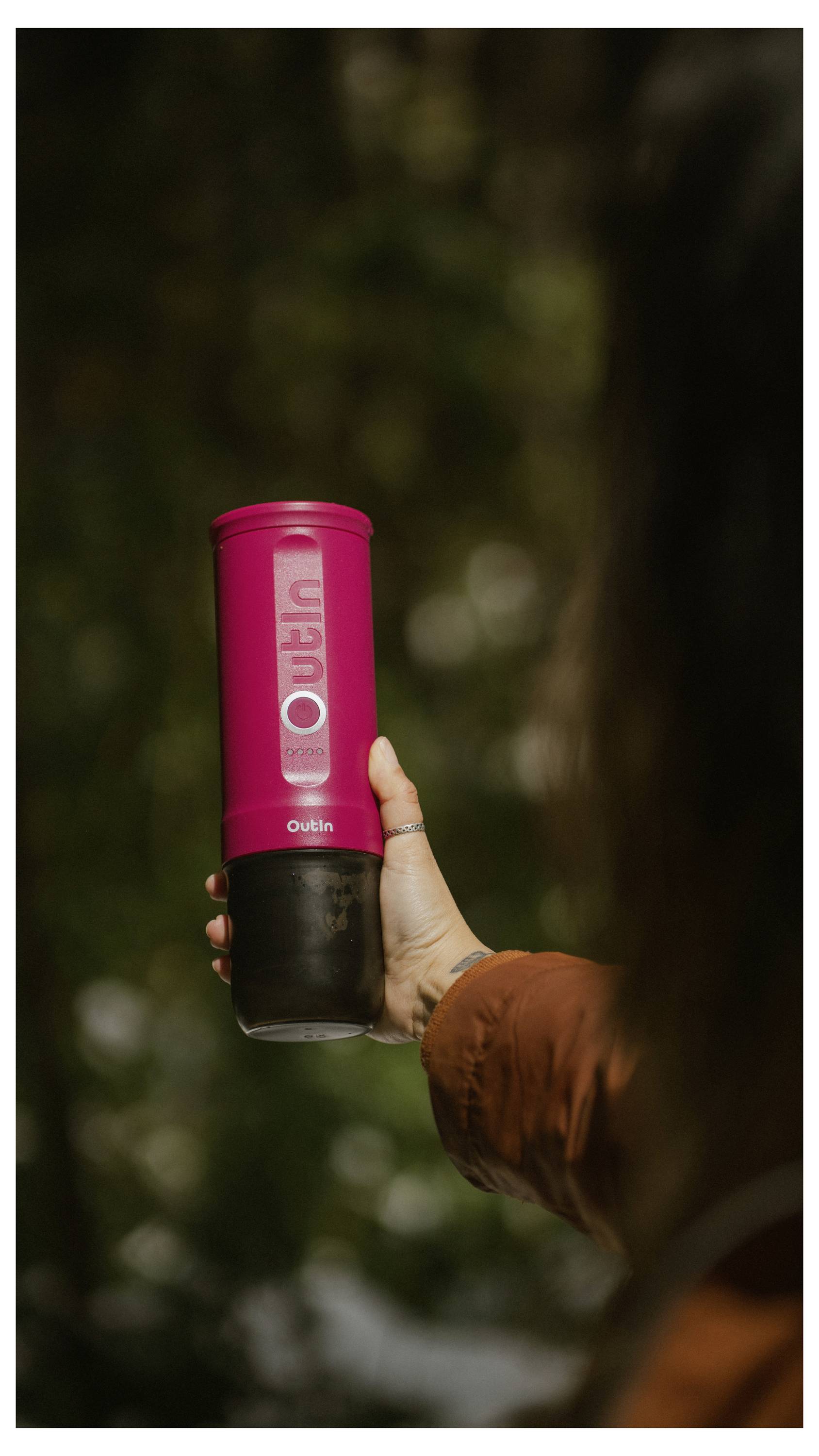 Eine Person, die eine rosa Reisebecher mit schwarzem Sockel hält, steht im Freien vor einem unscharfen grünen Hintergrund.