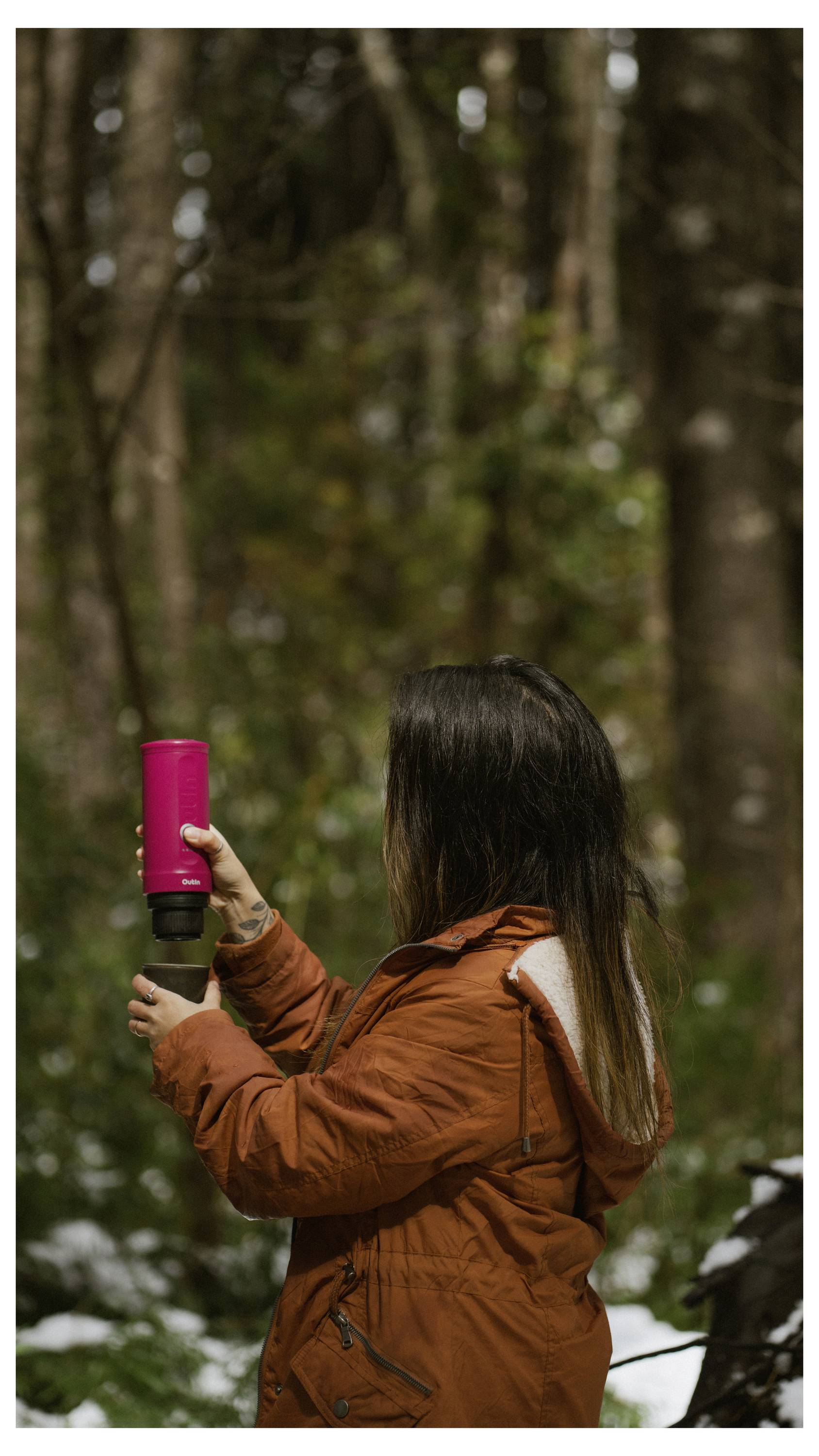 Eine Person in einer orangefarbenen Jacke hält eine rosa Thermoskanne in einer Waldumgebung mit unscharfen Bäumen im Hintergrund.