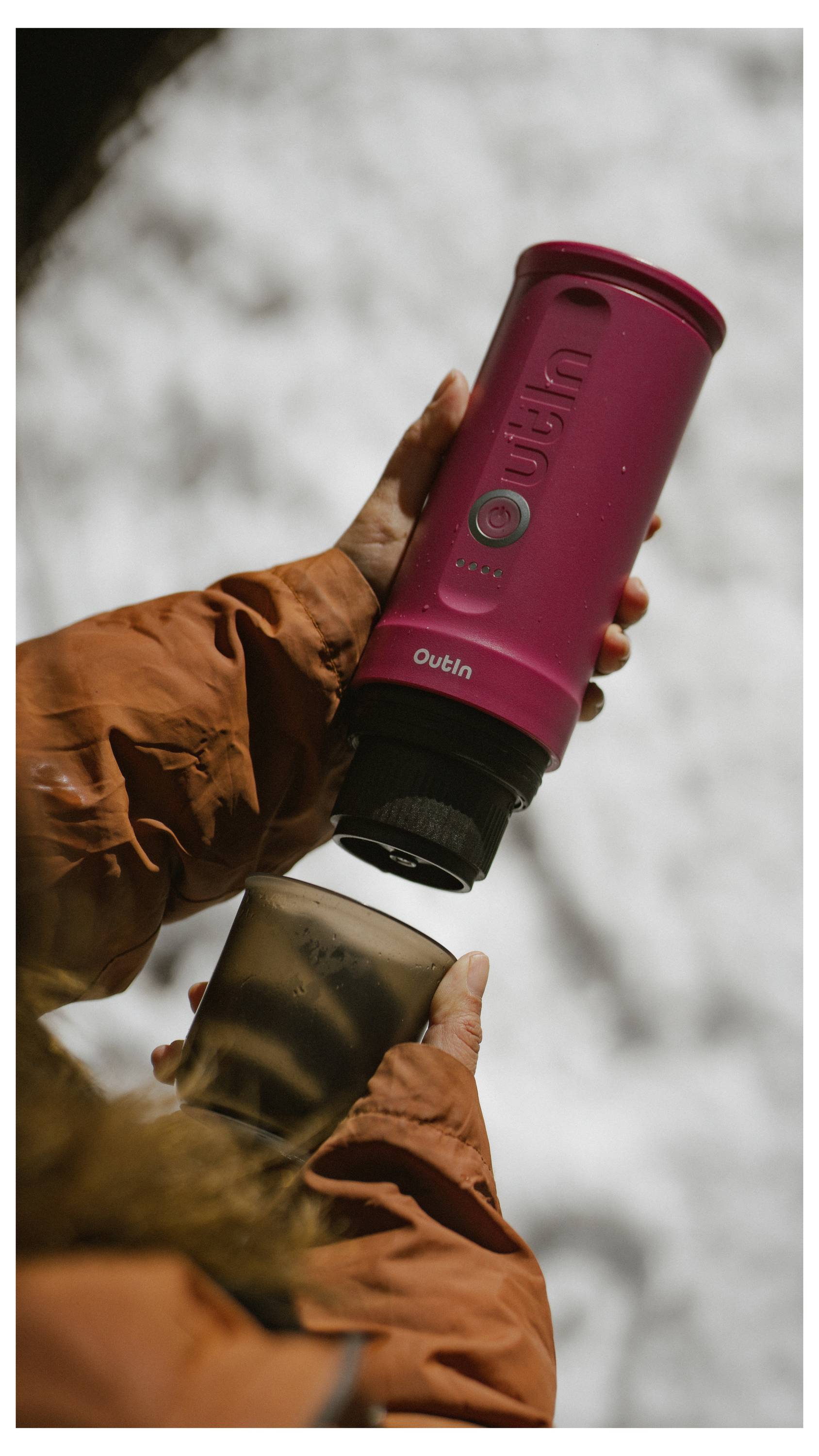Eine Person in einem orangefarbenen Wintermantel gießt Kaffee aus einer rosa tragbaren Kaffeemaschine in eine metallische Tasse, mit verschneitem Hintergrund.