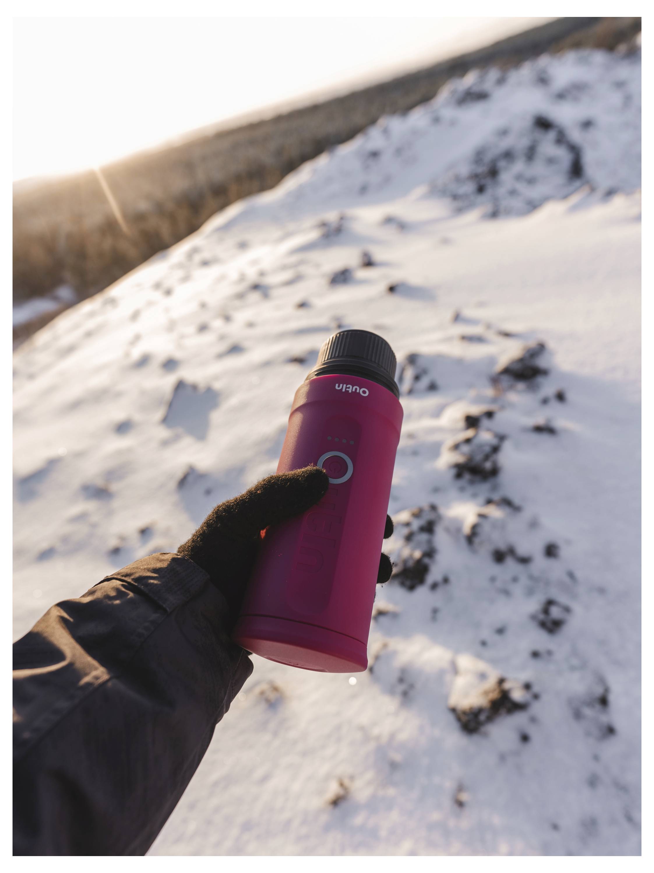 Eine behandschuhte Hand hält eine rote Thermoskanne vor einer schneebedeckten Bergkulisse bei Sonnenuntergang und deutet auf ein warmes Getränk während einer Winterwanderung hin.