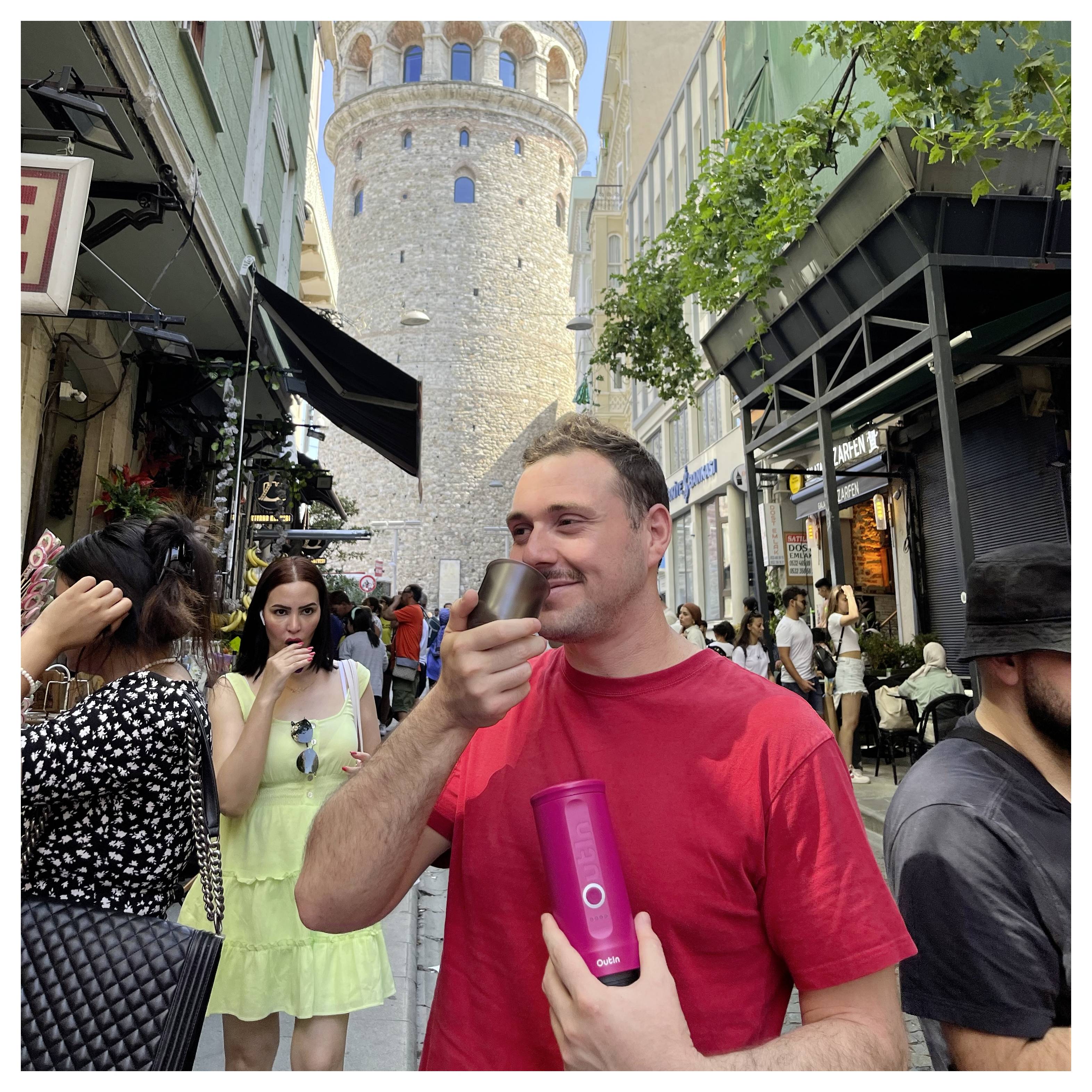 Eine Person in einem roten Hemd trinkt aus einer kleinen Tasse auf einer belebten Straße mit einem historischen Turm im Hintergrund; Menschen gehen in der Nähe vorbei.