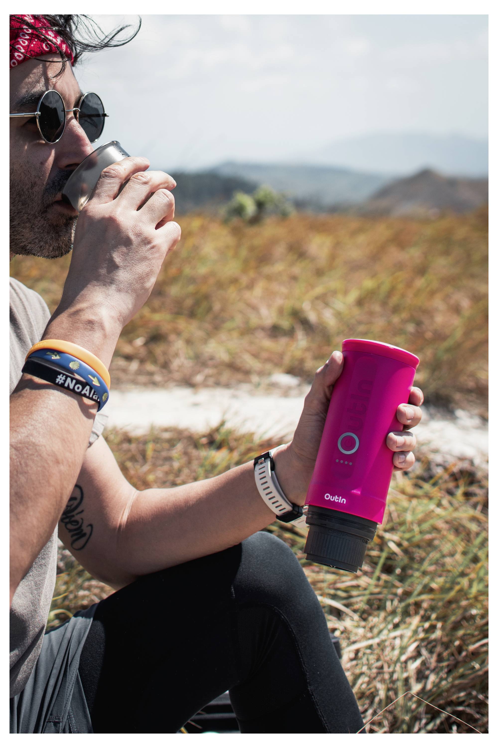 Eine Person im Freien auf einem Feld hält eine leuchtend rosa tragbare Kaffeemaschine mit der Marke "Qubik", während sie aus einer Metallkasse trinkt.