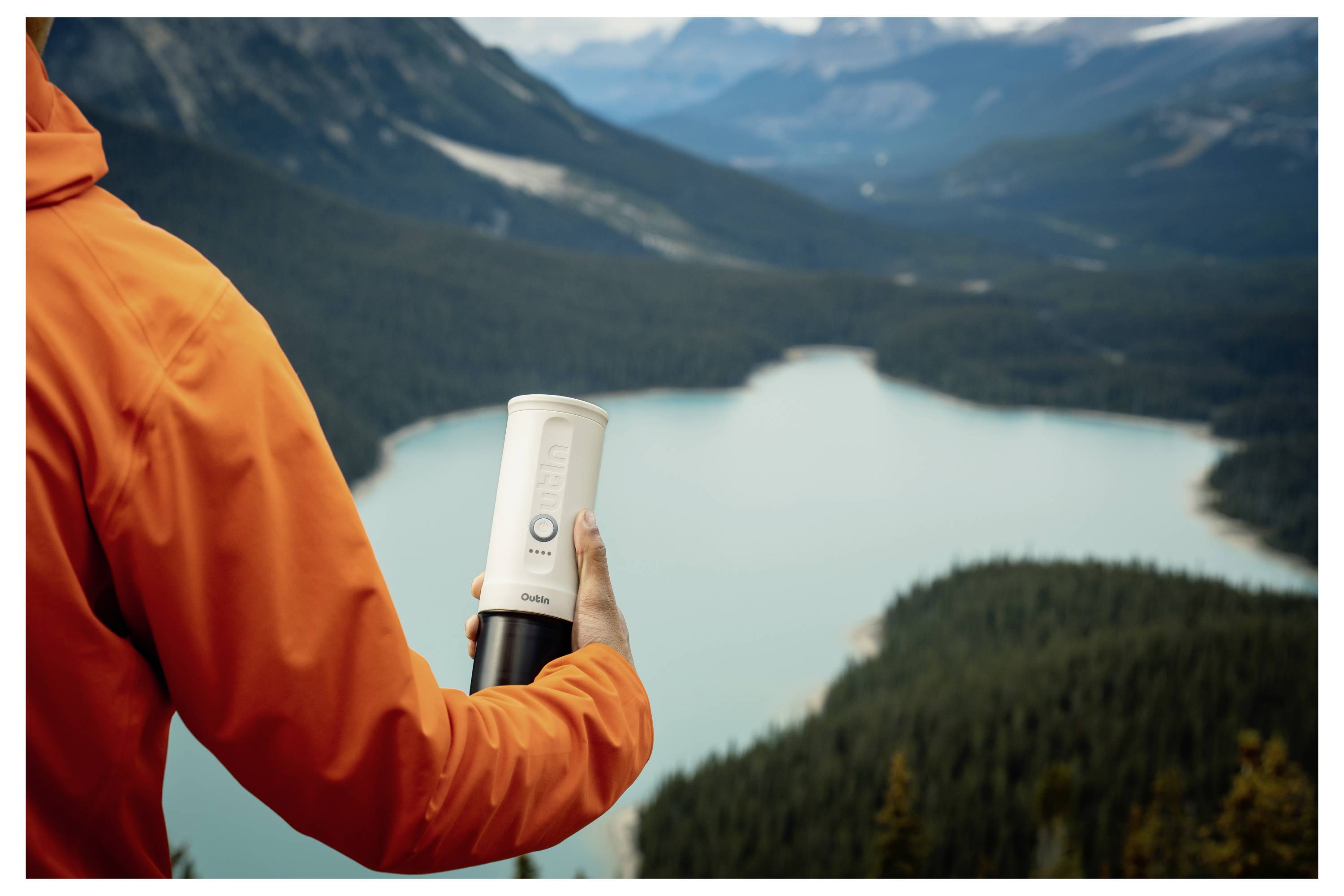 Eine Person in einer orangefarbenen Jacke hält ein weißes Gerät und überblickt einen malerischen Bergsee, was auf einen Wasserqualitätstest in der Natur hindeutet.