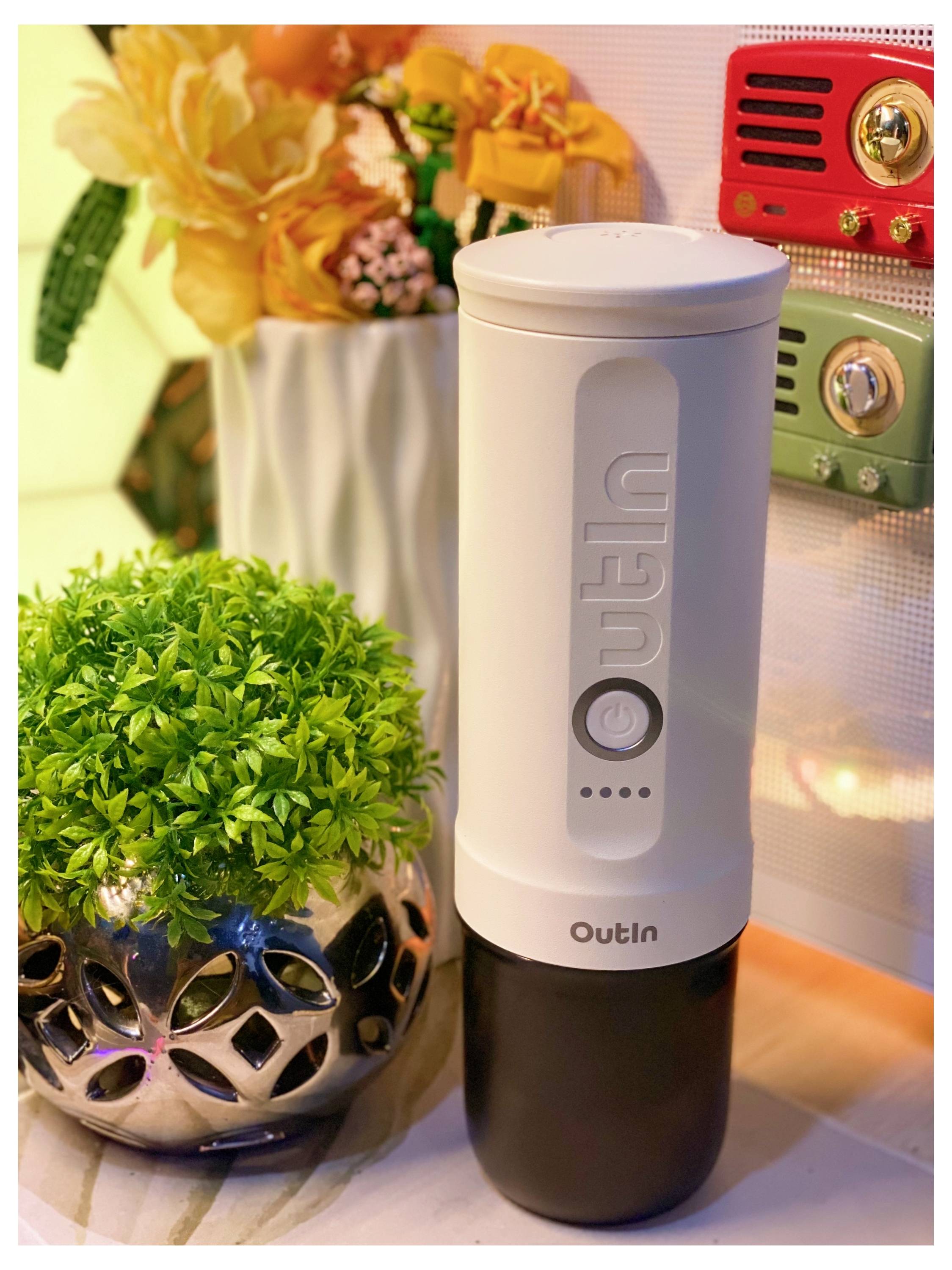 Ein weißer tragbarer Mixer mit der Aufschrift „Outln" neben einer Topfpflanze auf einer Arbeitsplatte, mit dekorativen Blumen und einem Retro-Radio im Hintergrund.