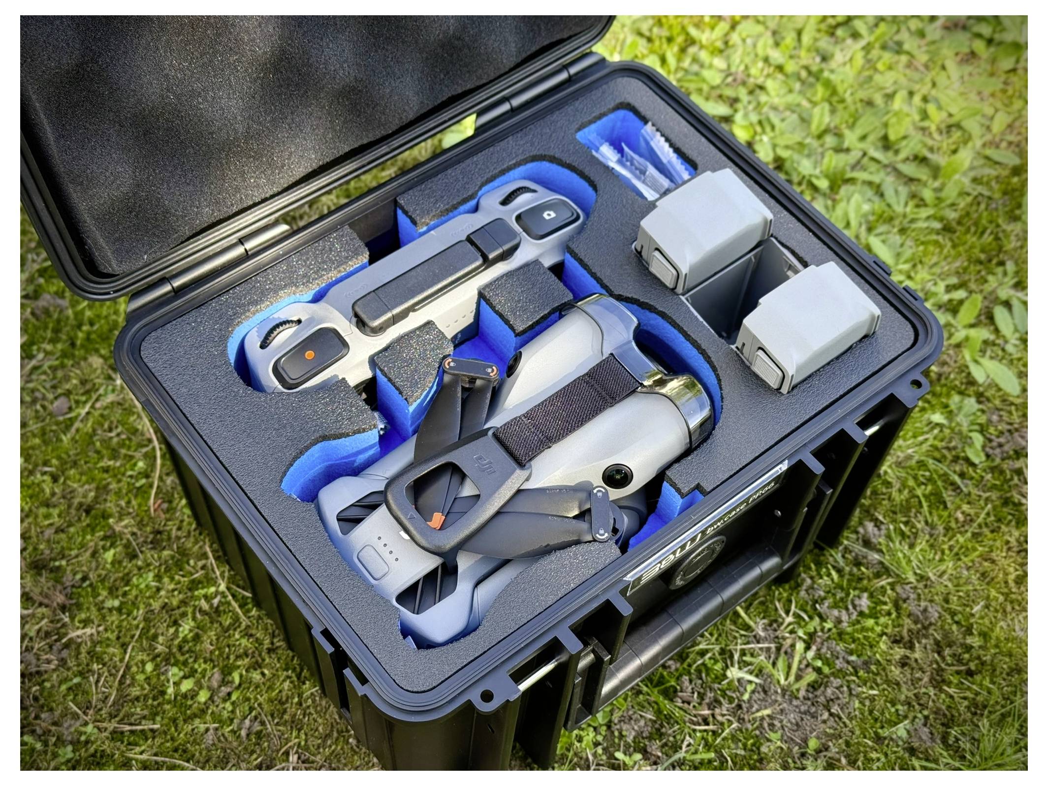 Ein offener Koffer auf Gras mit einer Drohne, einer Fernbedienung, zwei Batterien und Zubehör, organisiert in Schaumstoff-Fächern.