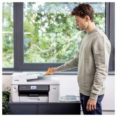 Ein Mann bedient einen Drucker und führt oranges Papier in den Scanner ein. Die Szene spielt sich in einem hellen Raum mit einem großen Fenster und Grünflächen außerhalb ab.