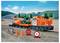 Ein Spielzeugzug mit einer orangefarbenen Lokomotive, einem Güterwagen mit Fahrzeug und einem Kesselwagen auf einem Gleis. Kegel, Stoppschild und Steine in der Nähe.