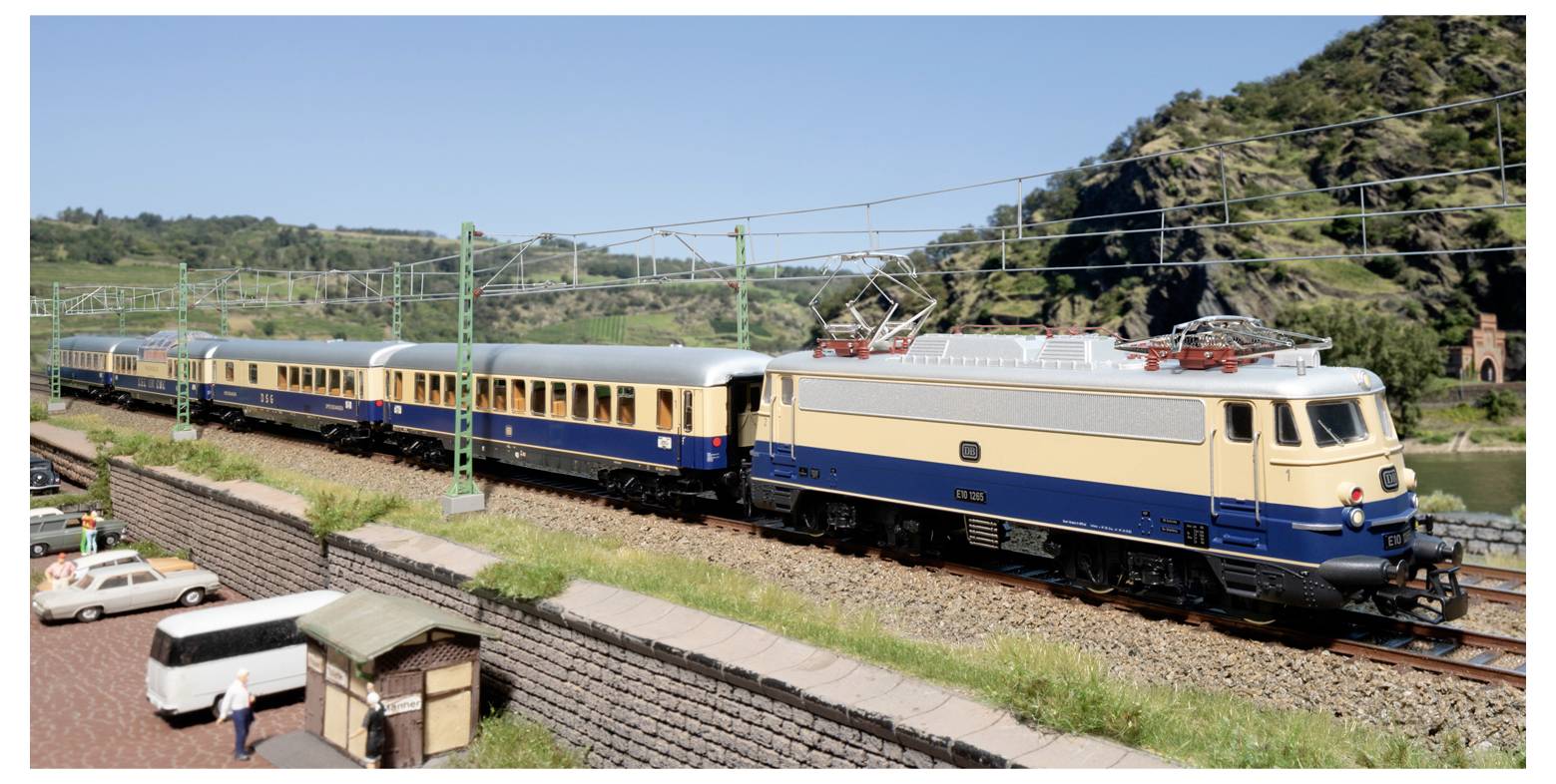 Ein Modelleisenbahn-Zug auf einer Spur fährt an einem malerischen Hintergrund mit Hügeln und Bäumen vorbei. Ein kleines Auto und eine Person sind im Vordergrund sichtbar.