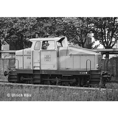 Eine vintage Diesellokomotive auf Gleisen, mit einem sichtbaren Lokführer in der Kabine. Bäume und Industriegebäude sind im Hintergrund.