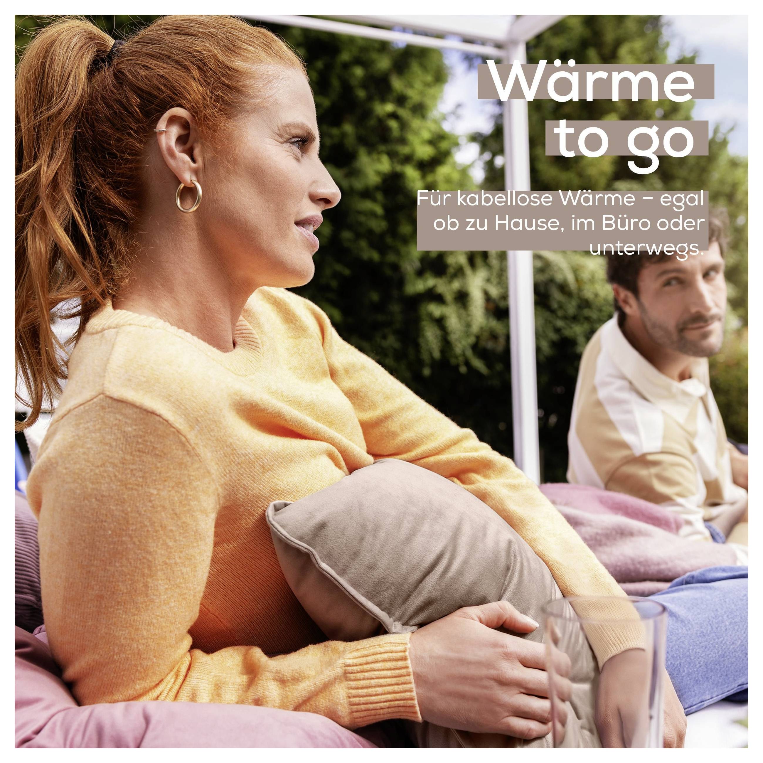 Eine Frau in einem orangefarbenen Pullover sitzt im Freien und hält ein Kissen, mit einem Mann im Hintergrund. Der Text lautet „Wärme to go".