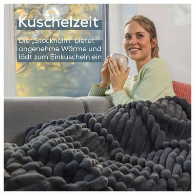 Eine Person sitzt in einer gemütlichen Umgebung auf einem Sofa, eingewickelt in eine dicke, strukturierte Decke und hält eine Tasse, mit großen Fenstern im Hintergrund.