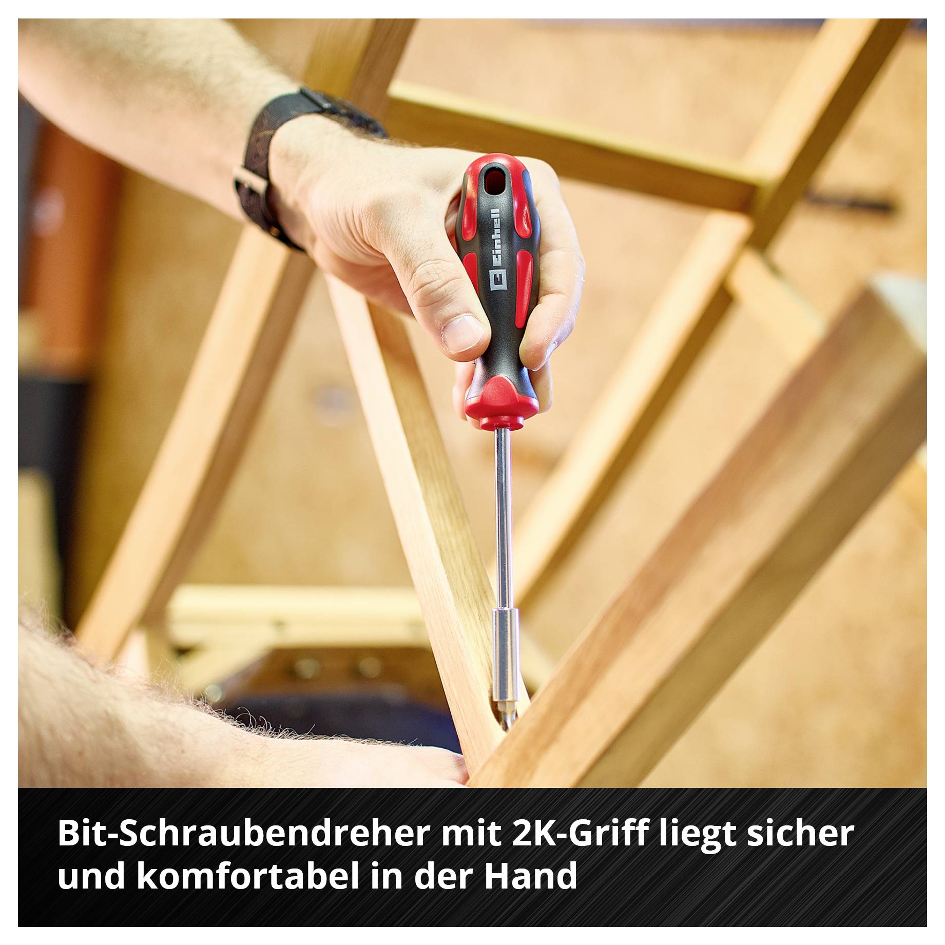 Ein Bit-Schraubendreher mit 2K-Griff wird benutzt, um eine Schraube in Holz zu drehen. Text: 'Bit-Schraubendreher mit 2K-Griff liegt sicher und komfortabel in der Hand'.