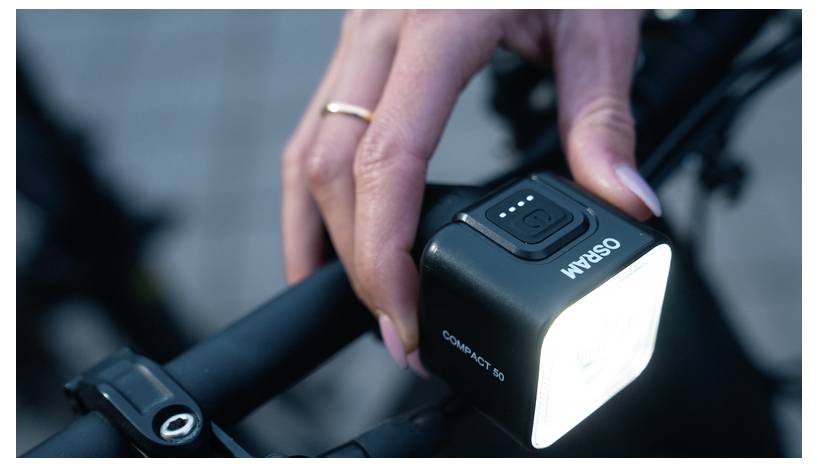 Eine Person stellt ein kompaktes Fahrradlicht ein, das an den Lenker montiert ist und den Bereich voraus beleuchtet. Das Gerät zeigt einen Leistungspegelanzeiger an.