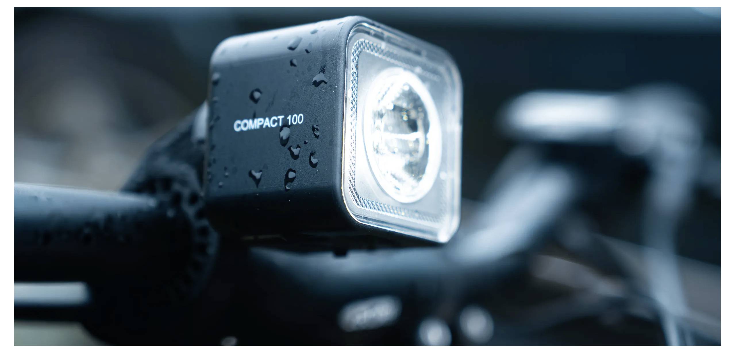 Eine Nahaufnahme eines Fahrradscheinwerfers mit der Bezeichnung „COMPACT 100" auf nassen Lenkerstangen, was auf die Verwendung bei regnerischen oder schlechten Lichtverhältnissen hindeutet.
