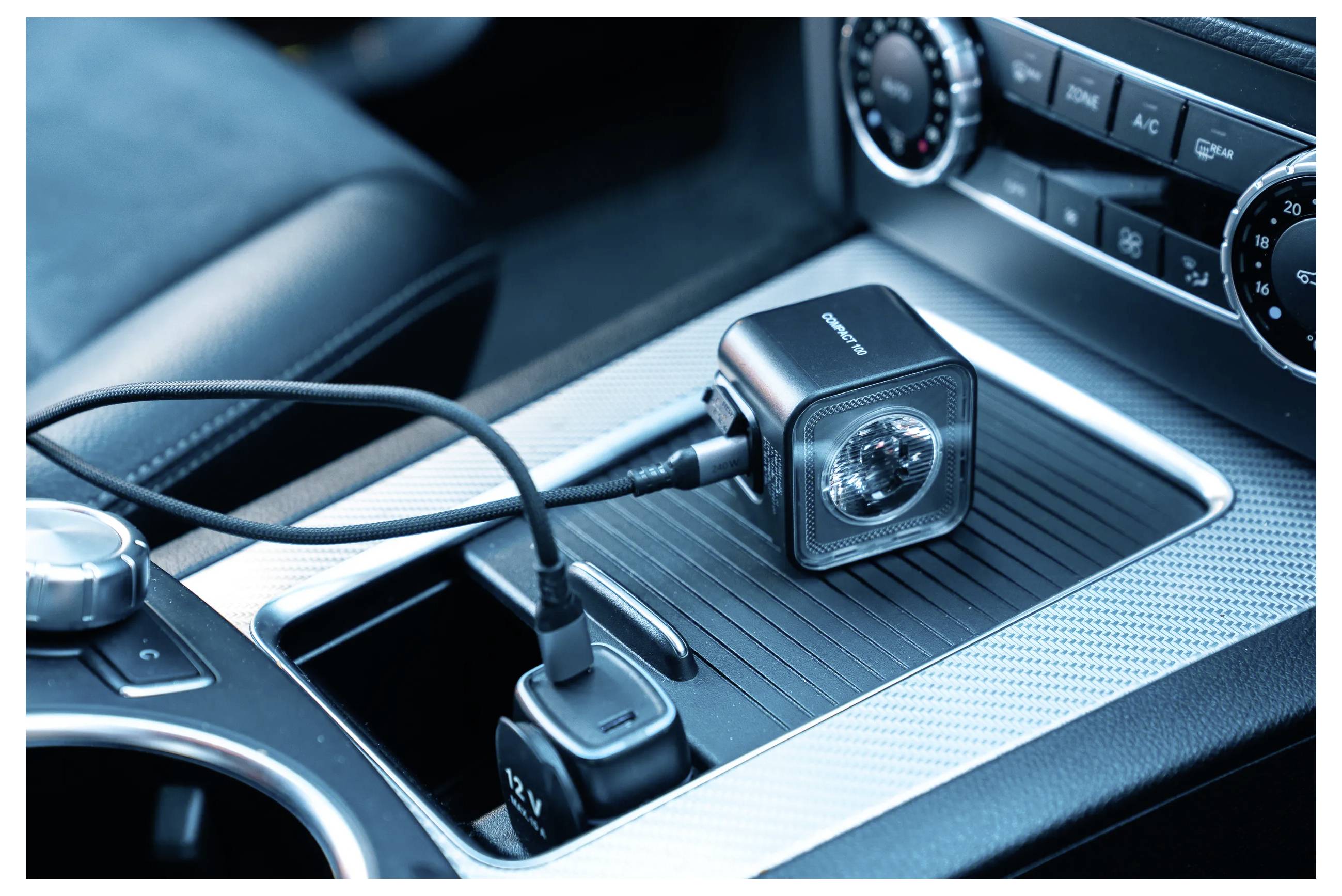 Armaturenbrett eines Autos mit dualem USB-Ladegerät, das in den Zigarettenanzünder eingesteckt ist und einen kleinen tragbaren Ventilator mit Strom versorgt. Klimaanlage-Bedienelemente sind sichtbar.