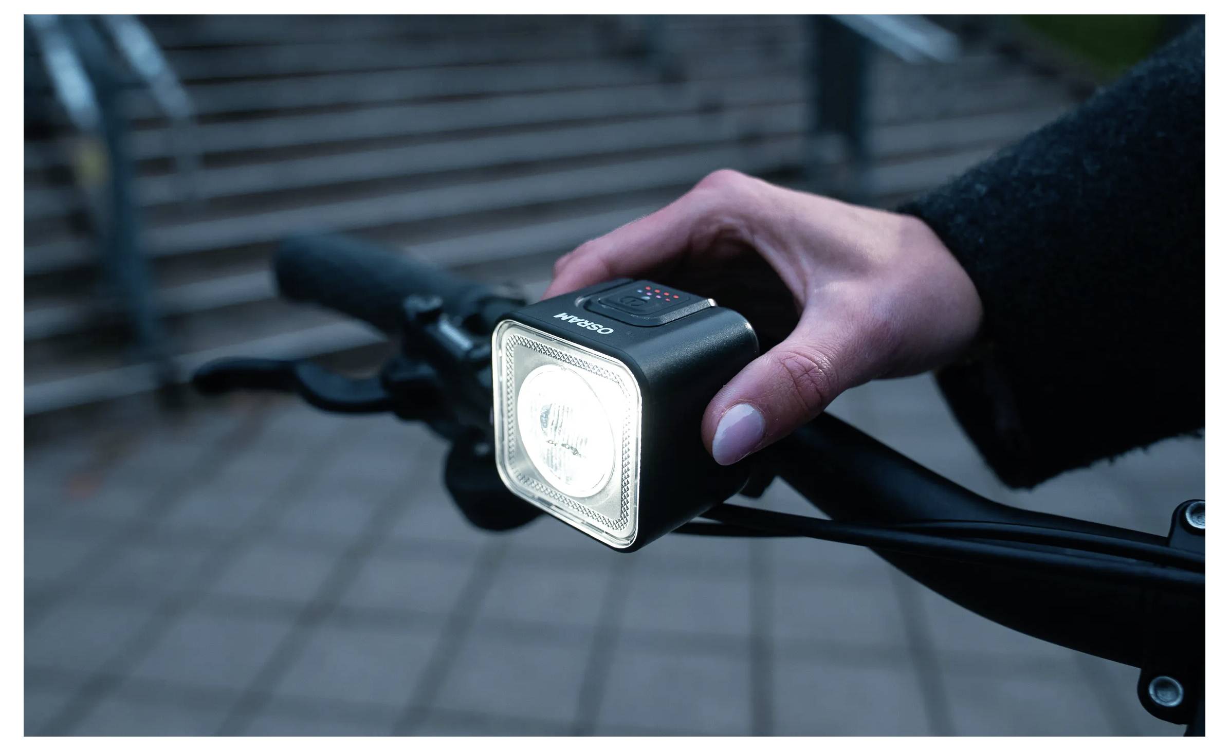 Eine Person schaltet ein helles Frontlicht an einem Fahrradlenker ein und bereitet sich auf eine Nachtfahrt vor. Der Hintergrund zeigt unscharfe Außentreppen.