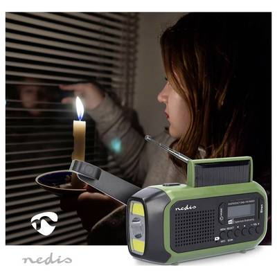 Eine Frau hält eine Kerze neben einem Fenster in der Dunkelheit. Im Vordergrund ist ein grünes Notfallradio zu sehen, das auf Vorsorgemaßnahmen hindeutet.