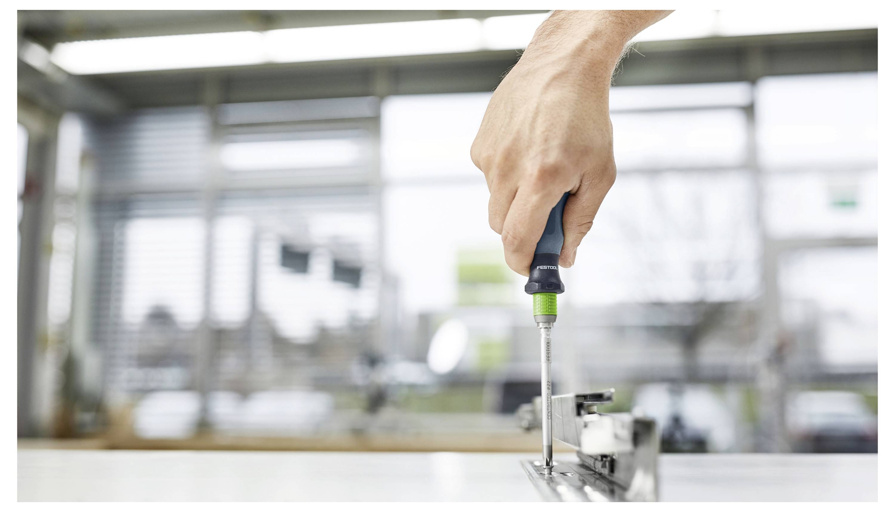 Eine Hand benutzt einen Schraubendreher, um ein Metallbeschlag-Teil in einer hellen, industriellen Umgebung zusammenzubauen oder einzustellen.
