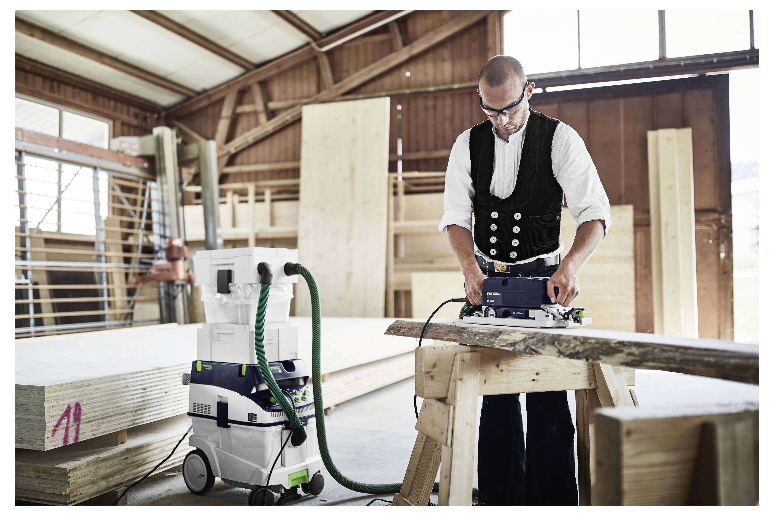 'Eine Werkstatt mit einer Festool-Holzbearbeitungsanlage, bestehend aus einer Tischkreissäge und einem Staubabsauggerät, in einer modernen, lichtdurchfluteten Umgebung.'