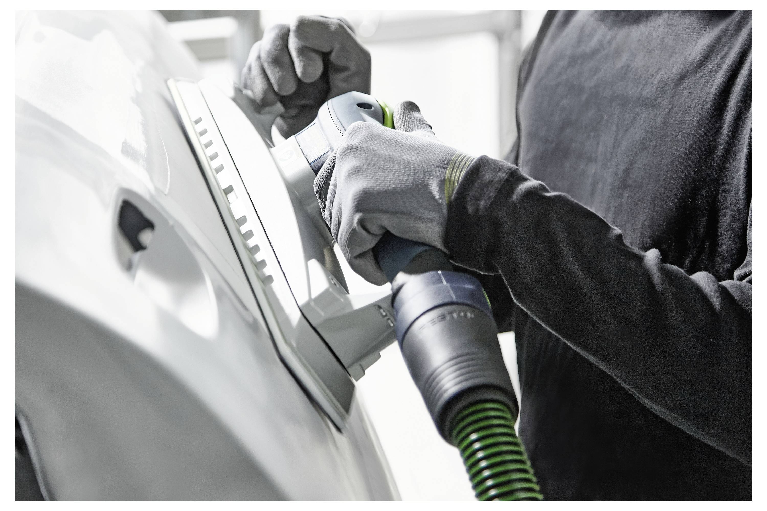 Eine Person trägt Handschuhe und verwendet einen Schleiffer auf der Karosserie eines Autos, um die Oberfläche in einer Autowerkstatt zu glätten.