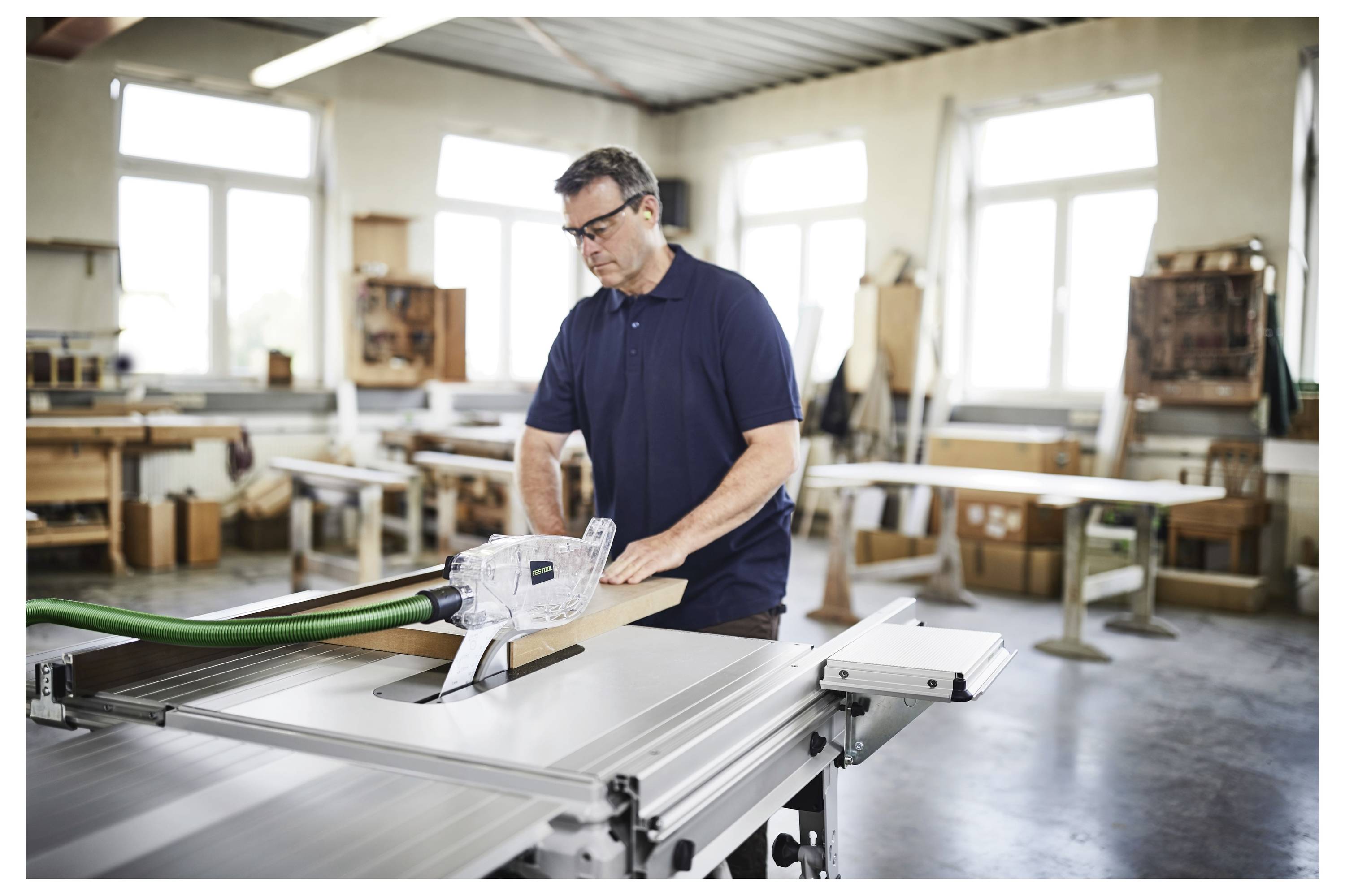 Ein Mann in einer Werkstatt bedient eine Tischkreissäge und schneidet Holz. Der Raum ist hell mit großen Fenstern und Regalen, die mit Werkzeugen und Materialien gefüllt sind.