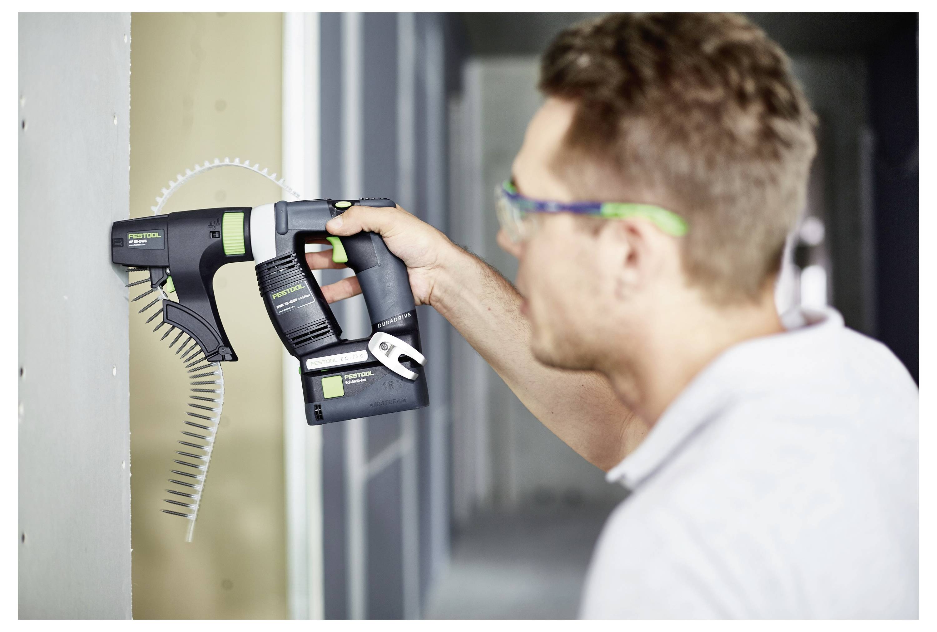 Eine Person benutzt einen kabellosen Elektroschrauber, um Schrauben in eine Wand zu treiben. Sie trägt eine Schutzbrille und wirkt konzentriert auf die Aufgabe.