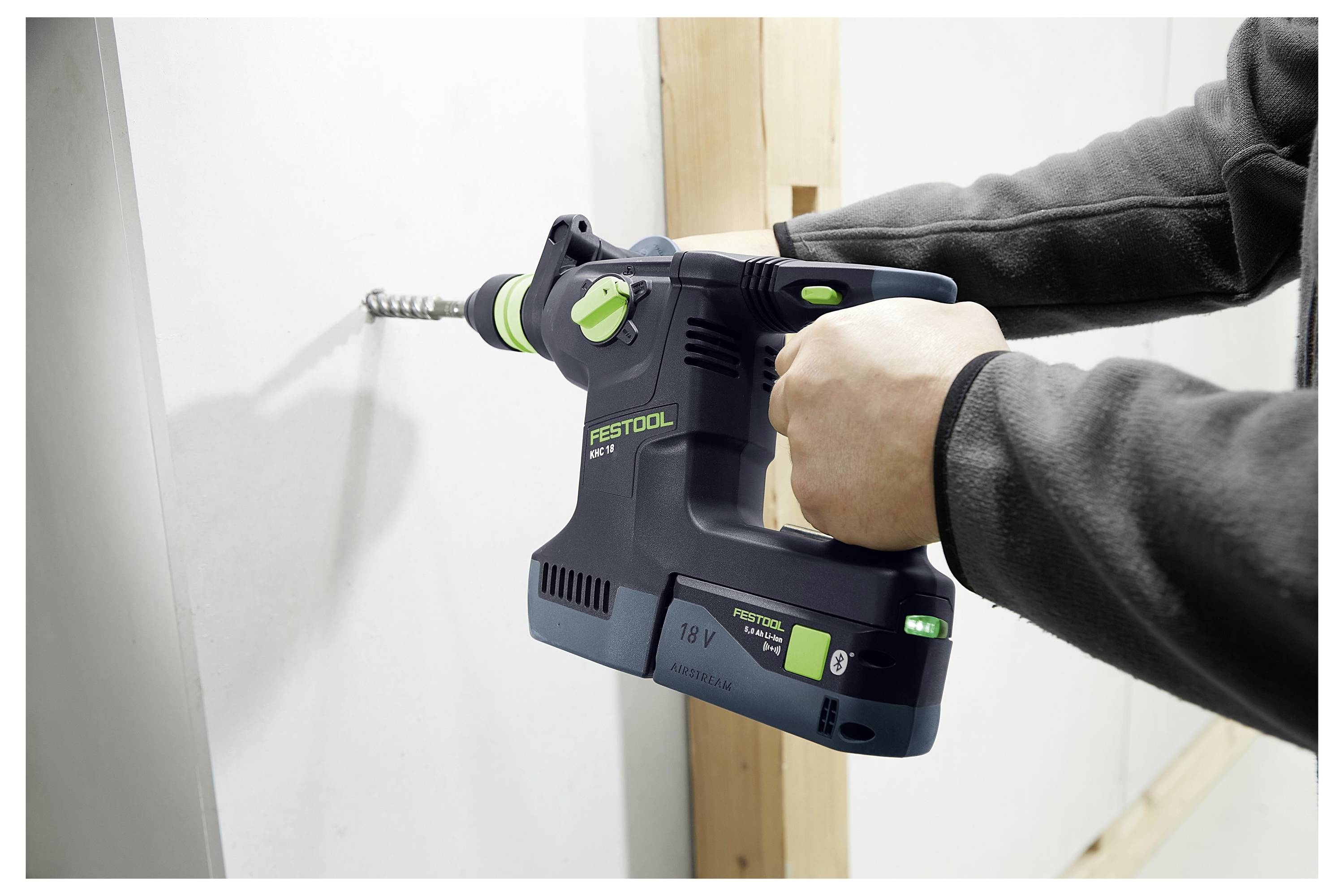Eine Person benutzt ein Elektrowerkzeug, um eine Wand zu schneiden, wobei sie sich auf Präzision und Kontrolle konzentriert. Das Werkzeug hat ein grün-schwarzes Design und einen Schlauch.