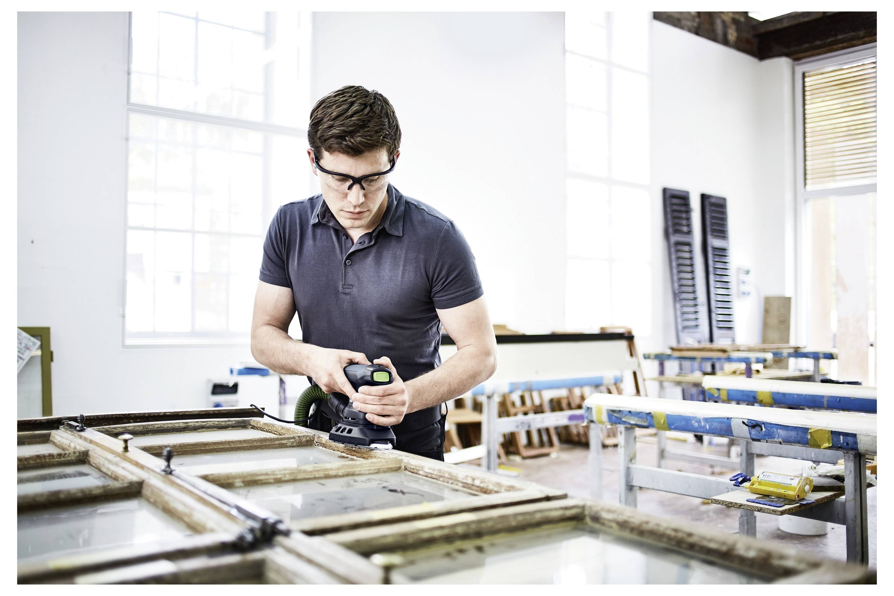 Eine Person mit einer Schutzbrille bedient ein Elektrowerkzeug, um einen Holzfensterrahmen in einer mit natürlichem Licht gefüllten Werkstatt zu überarbeiten.