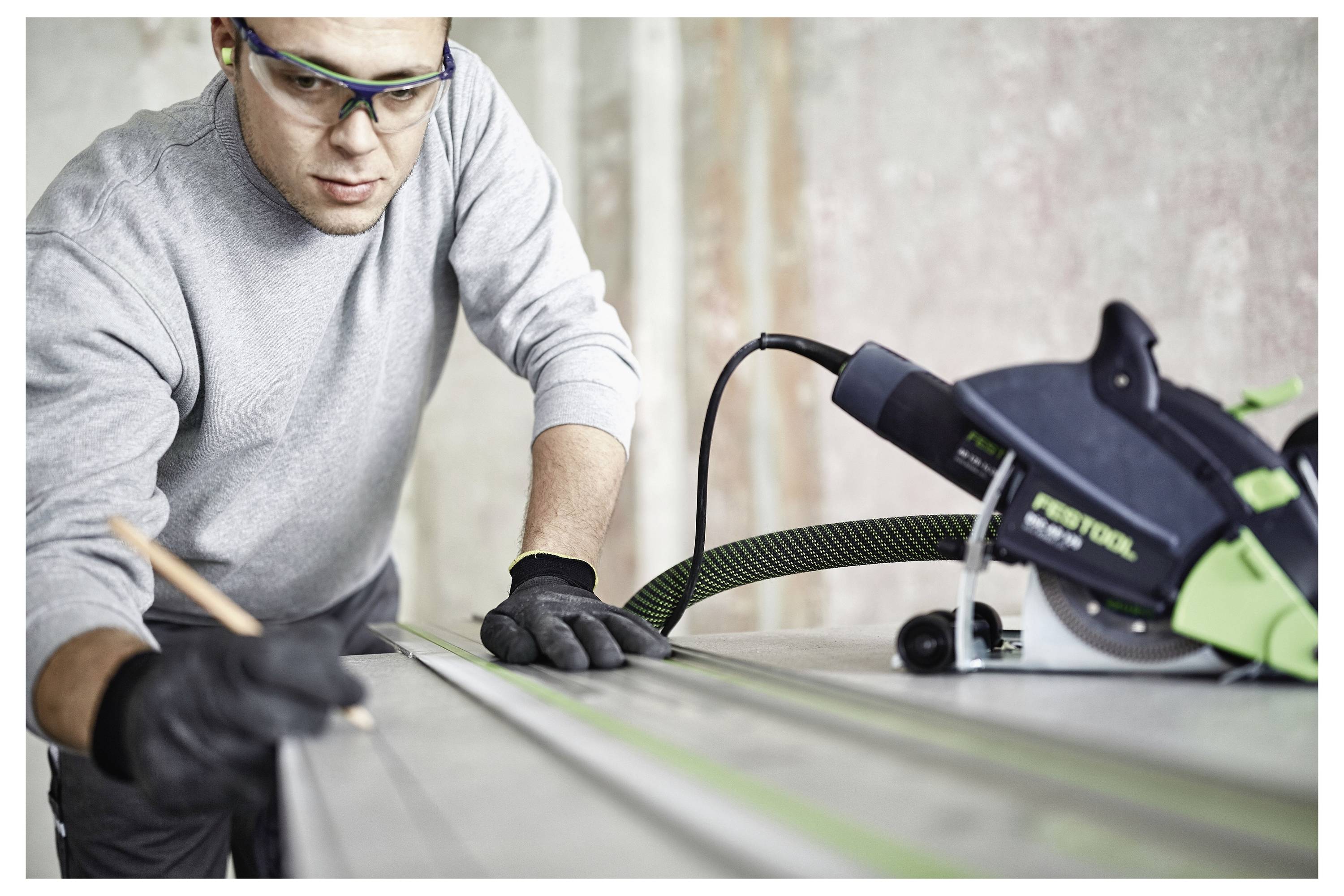 Eine Person, die eine Schutzbrille und Handschuhe trägt, misst sorgfältig ein Stück Holz mit einem Bleistift, während eine Elektrosäge in der Nähe auf dem Tisch liegt.
