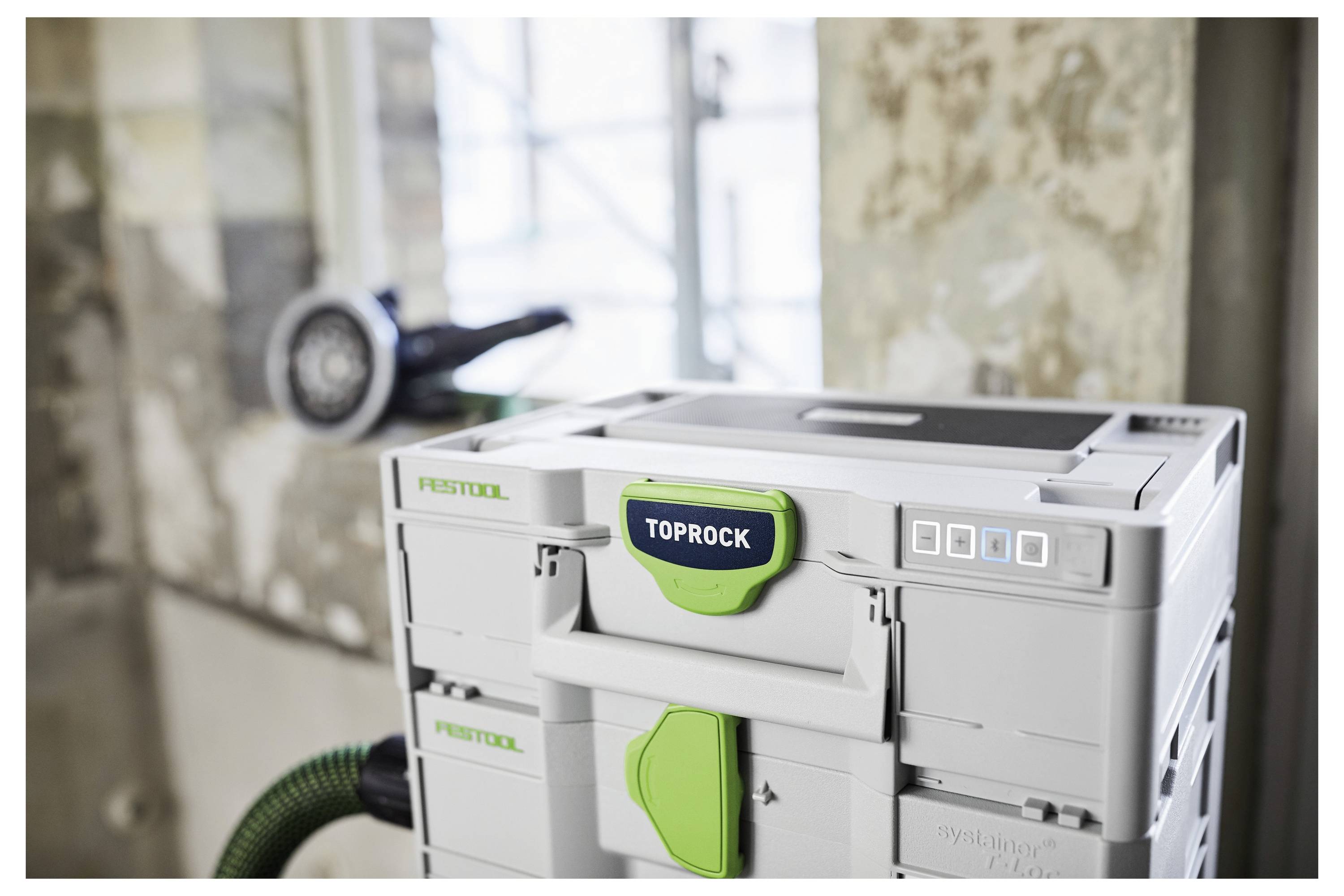 Eine graue Festool TOPROCK Werkzeugkiste mit grünen Akzenten steht in einer Werkstattsituation, wobei ein Werkzeug im Hintergrund an der Wand hängt.