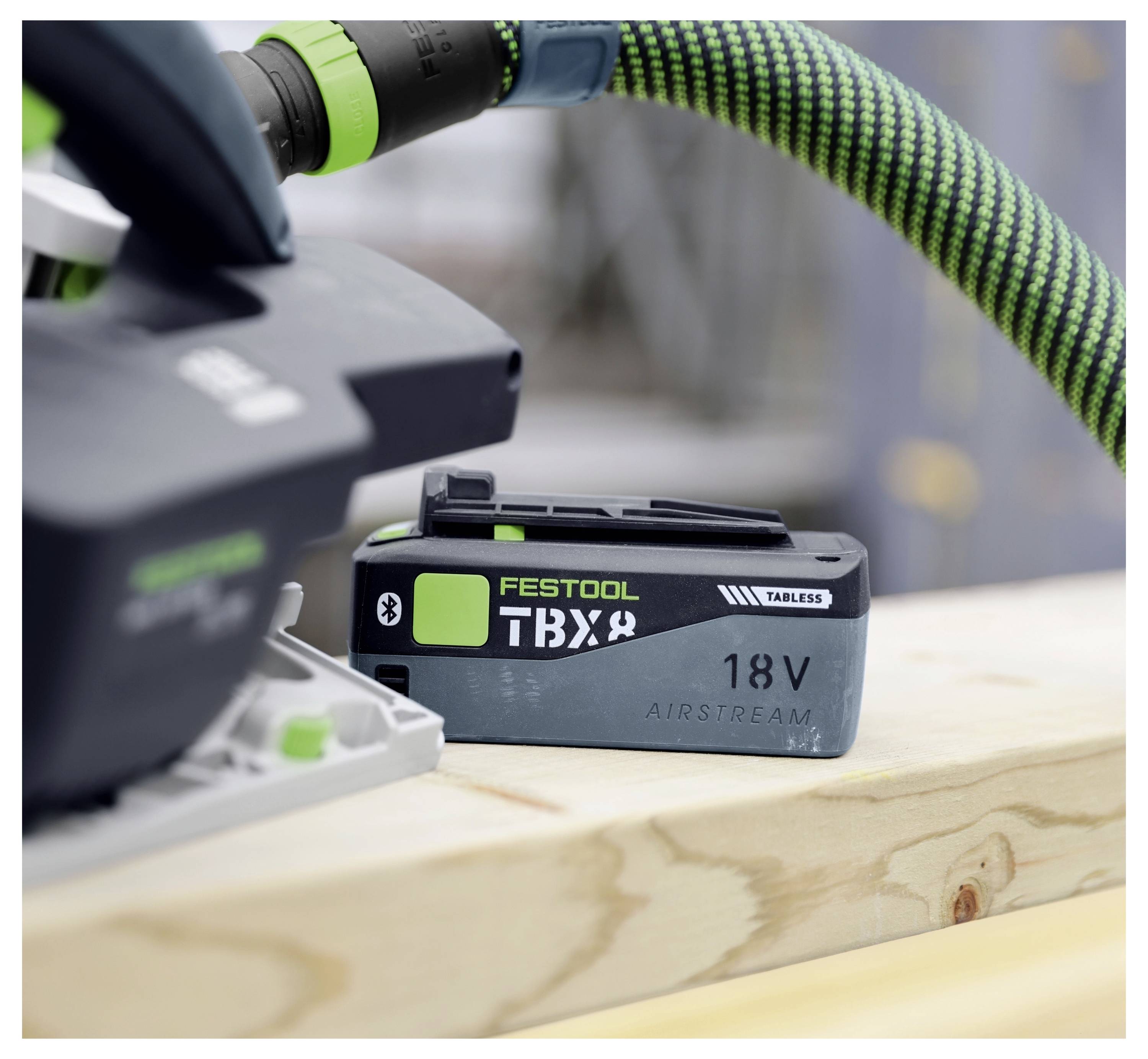 Eine Elektrowerkzeug-Batterie mit der Bezeichnung "Festool TBX 18V Airstream" ist an einem Werkzeug auf einer Holzoberfläche befestigt, wobei ein grüner Schlauch in der Nähe angeschlossen ist.