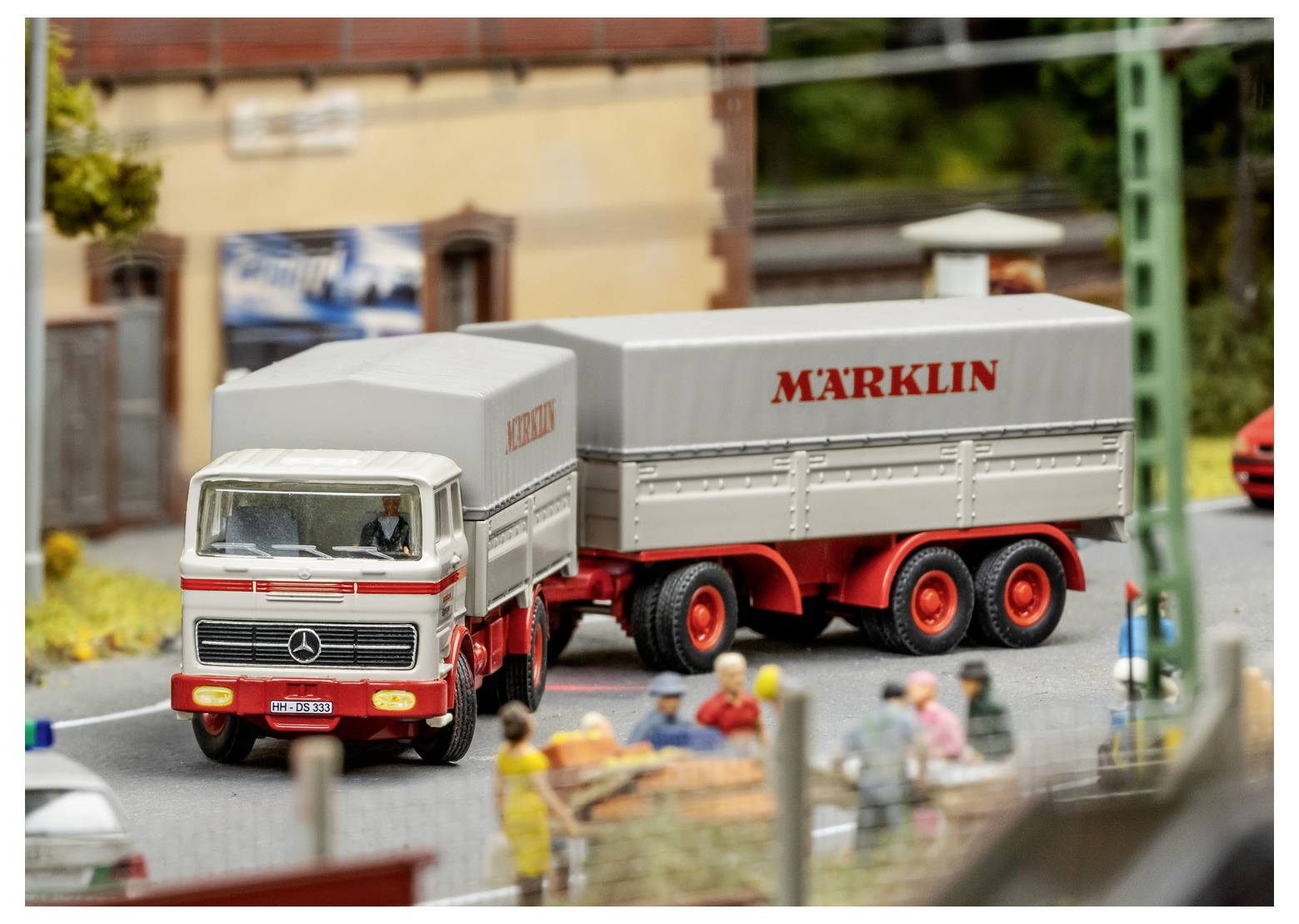 Eine Modellszene mit einem Vintage-Mercedes-Lastkraftwagen mit einem mit "Märklin" beschrifteten Anhänger, umgeben von Modellpersonen in einer Kleinstadt-Szenerie.