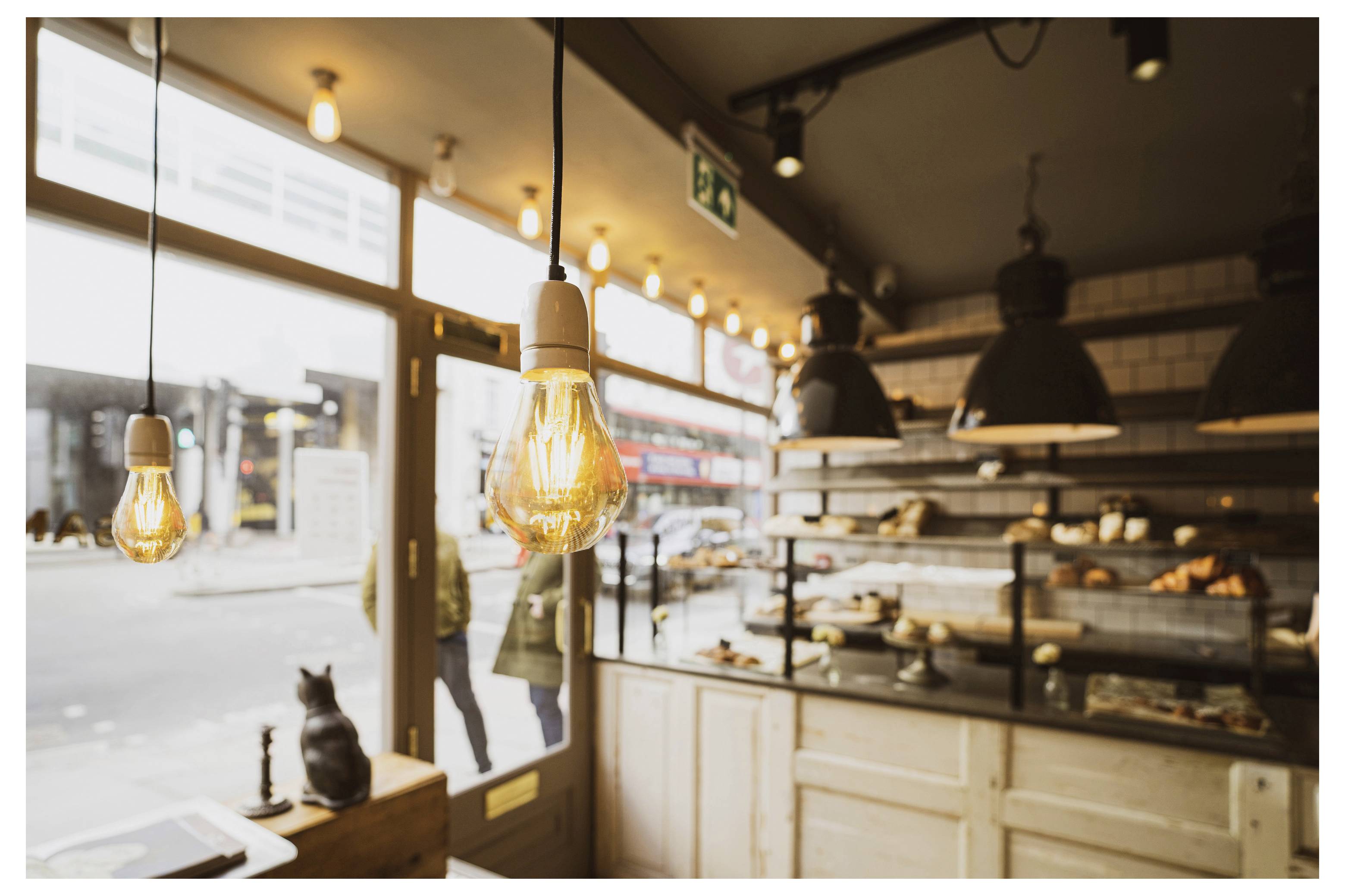 Inneres einer gemütlichen Bäckerei oder eines Cafés mit warmen hängenden Lichtern, einer Glasvitrine für Gebäck und einer Katze, die auf der Fensterbank am vorderen Fenster sitzt.